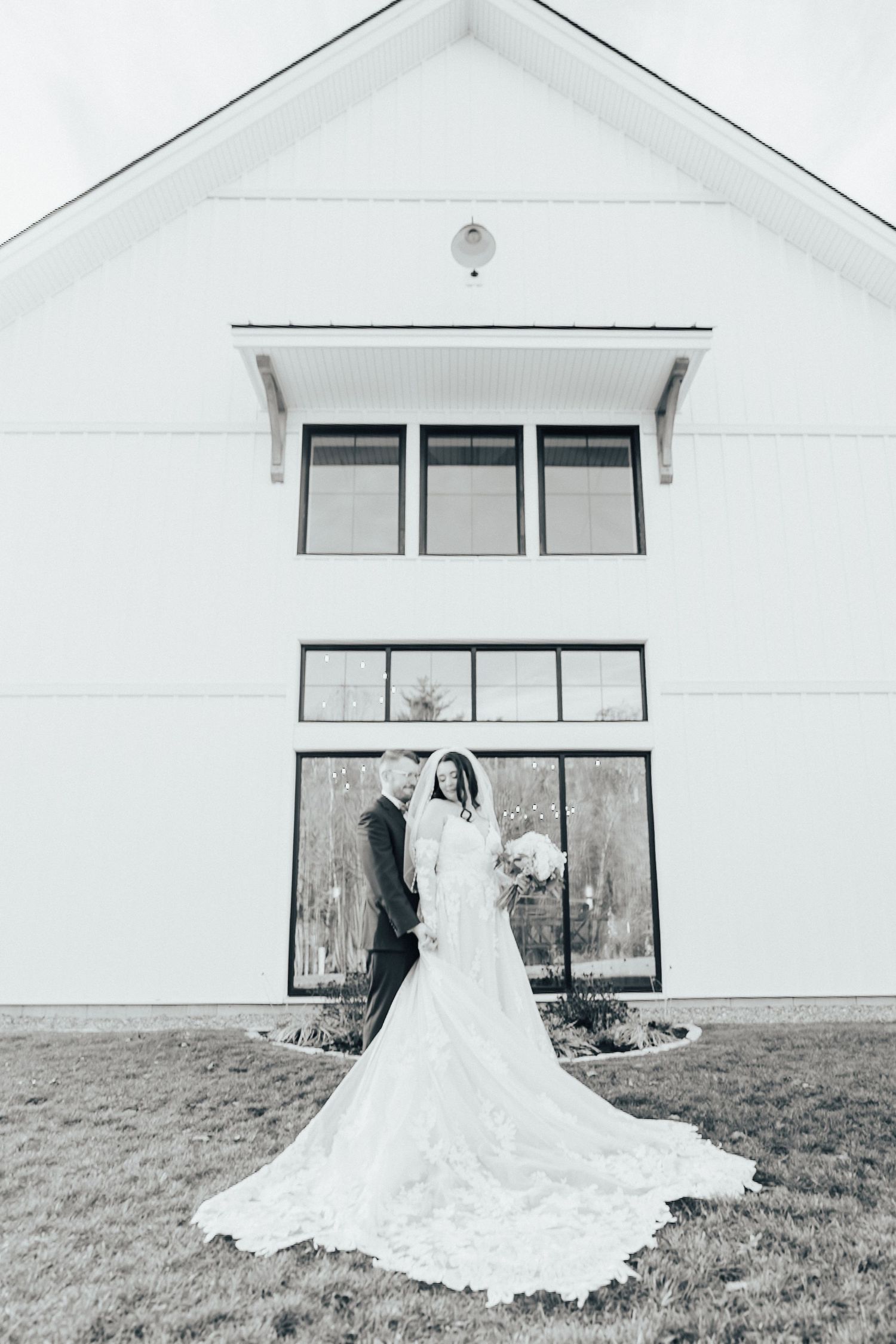 A bride in a flowing white wedding dress with a long train stands against a white building exterior.