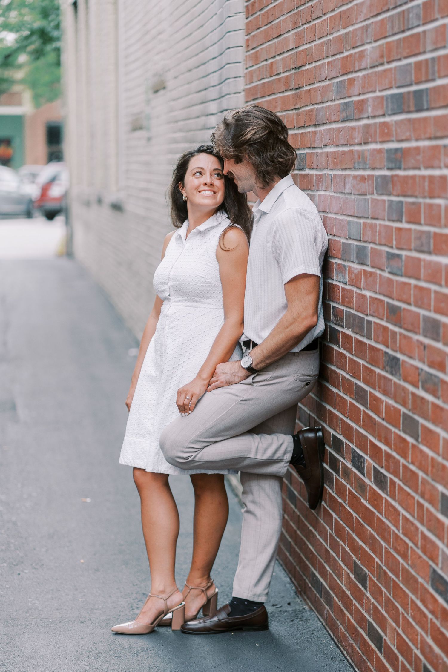 Couple in coordinated white and beige outfits posing against brick wall in urban setting.