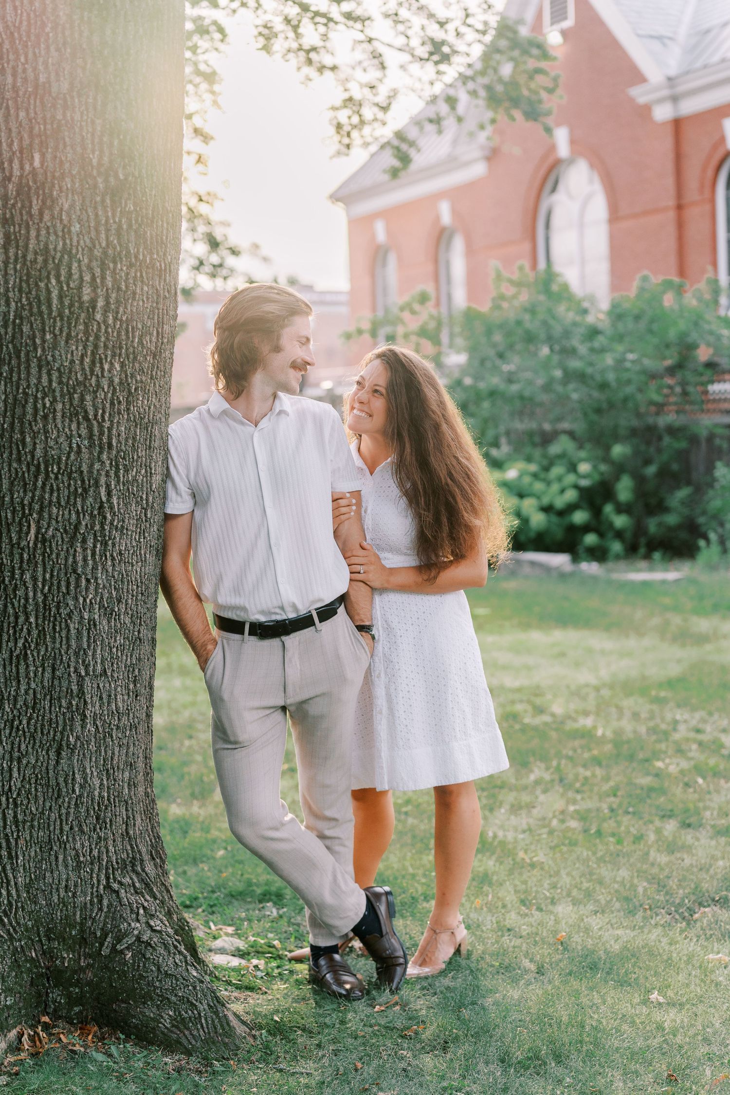 A couple in white outfits pose playfully near a large tree trunk on a summer day.