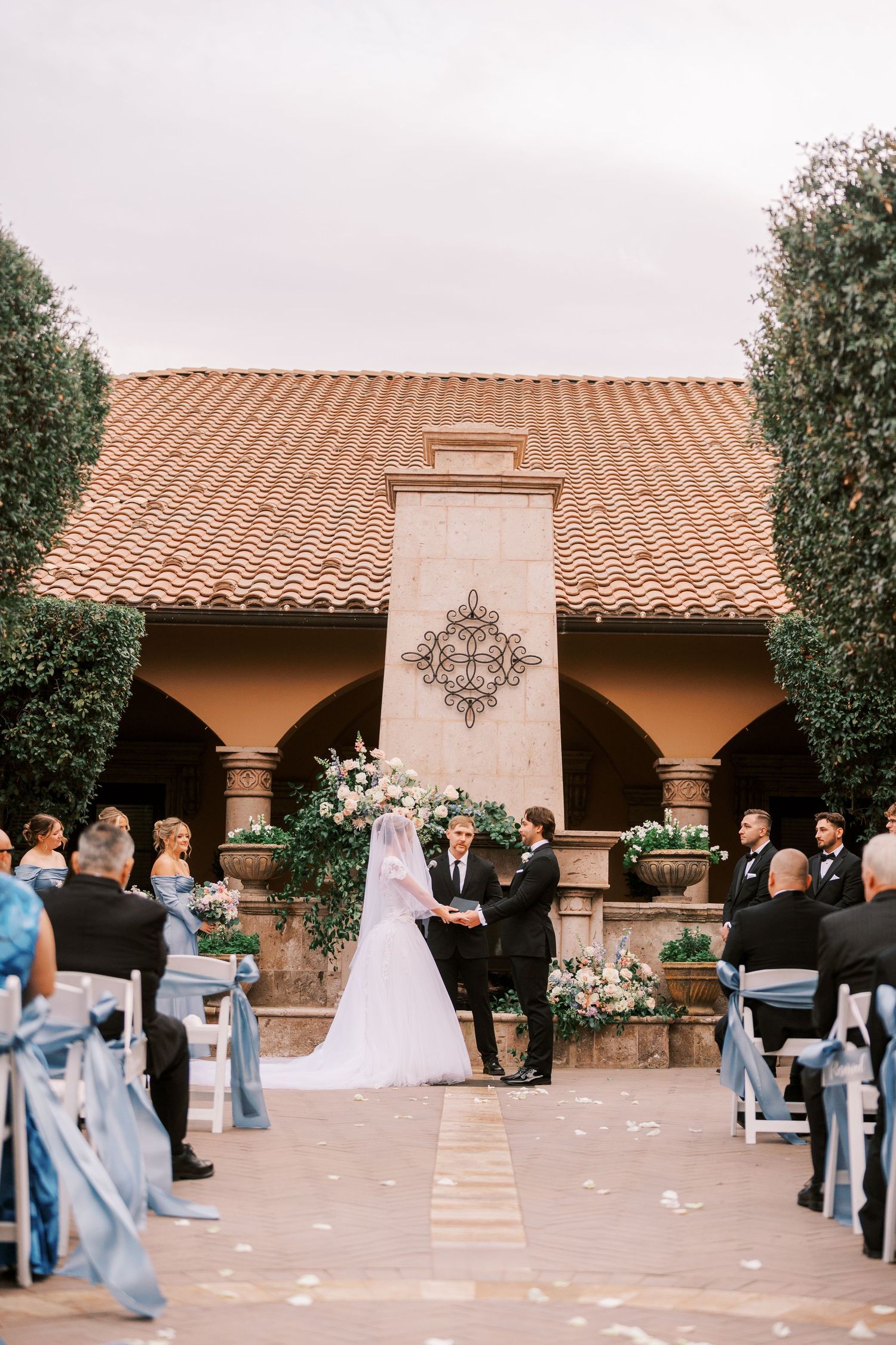 Outdoor wedding ceremony at Spanish-style venue with terra cotta roof and decorative columns, featuring light blue decor.