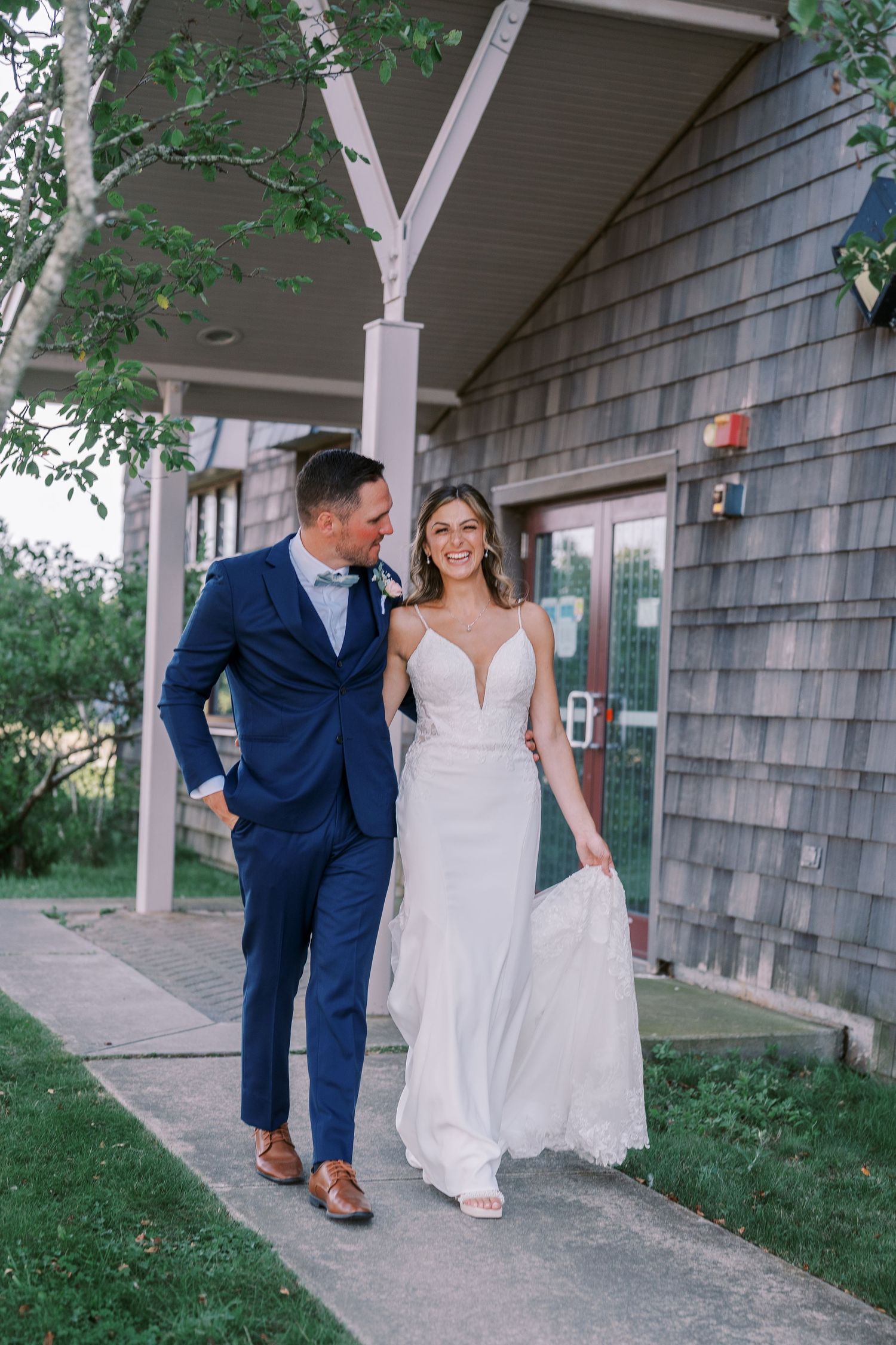 A bride in white dress and groom in navy suit share intimate moments along a rustic building exterior.