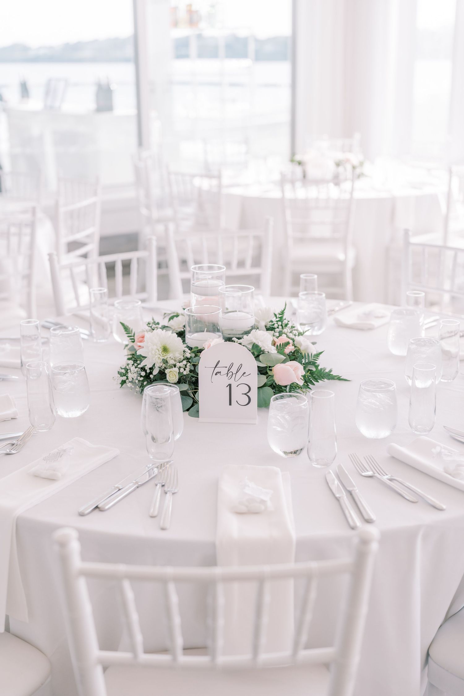 Elegant white wedding reception tables set with floral centerpieces and table numbers in a bright, airy venue.