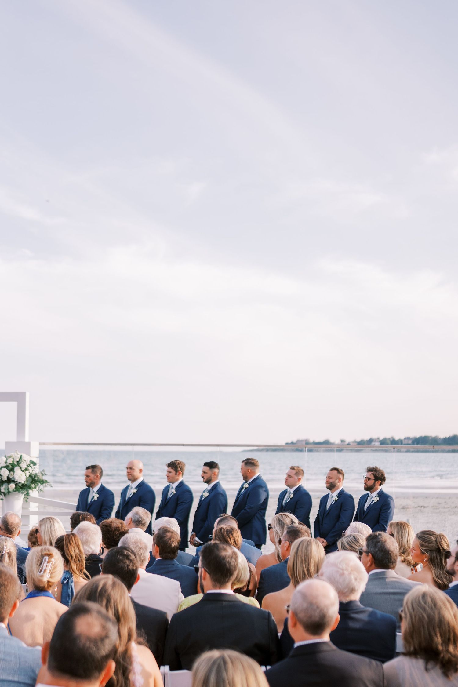 Wedding ceremony guests seated in rows facing the ocean as they watch a beachside service in progress.