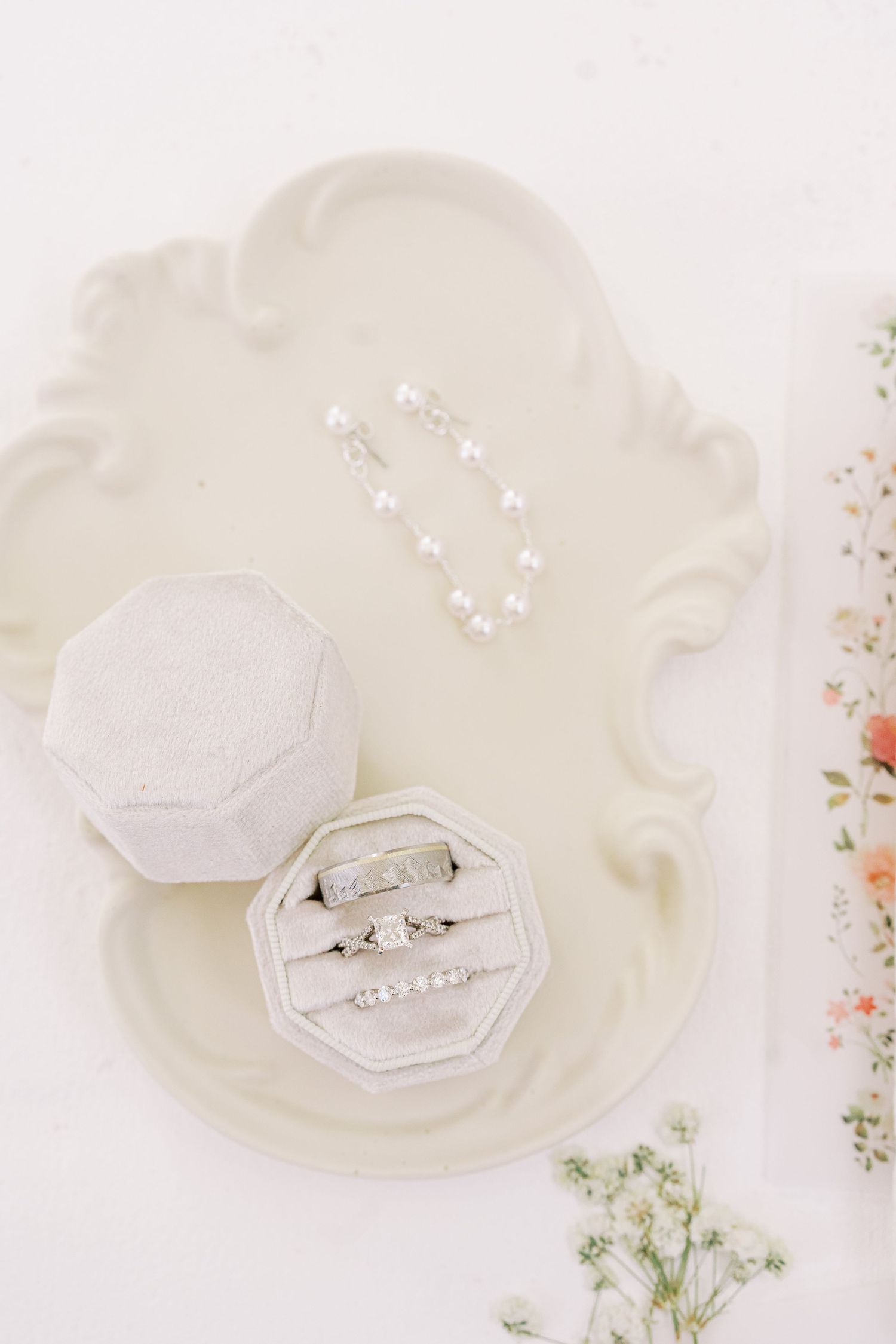 Wedding rings displayed in a white hexagonal ring box surrounded by delicate floral arrangements on a light background.