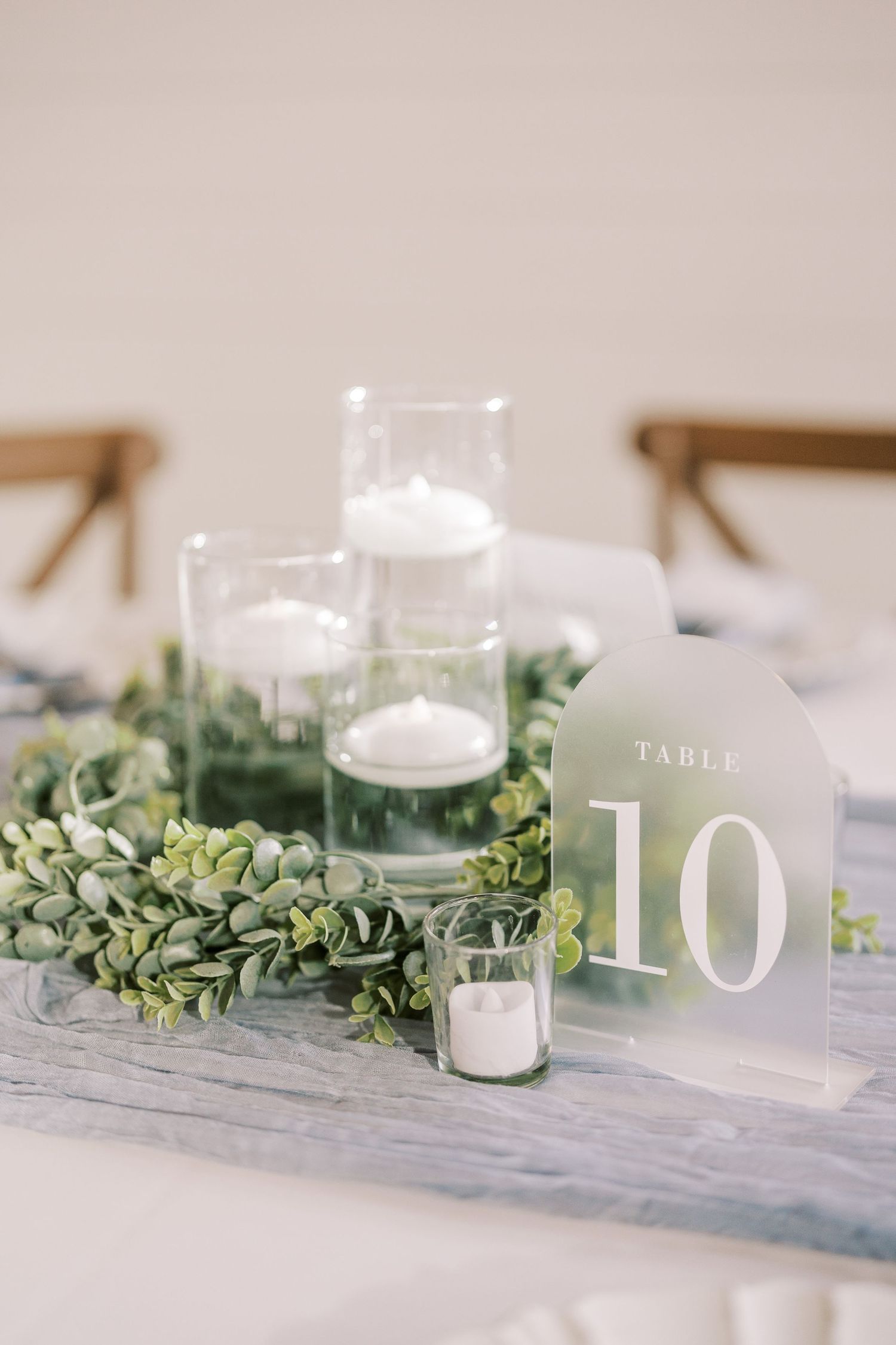 Wedding table centerpiece features eucalyptus garland, white candles and acrylic table number on gray linen.