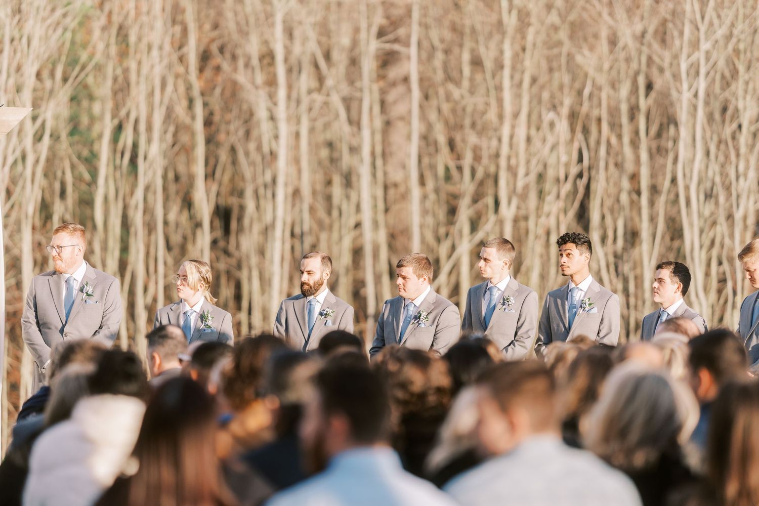 Wedding party poses together in front of a scenic wooded backdrop during an autumn outdoor celebration.