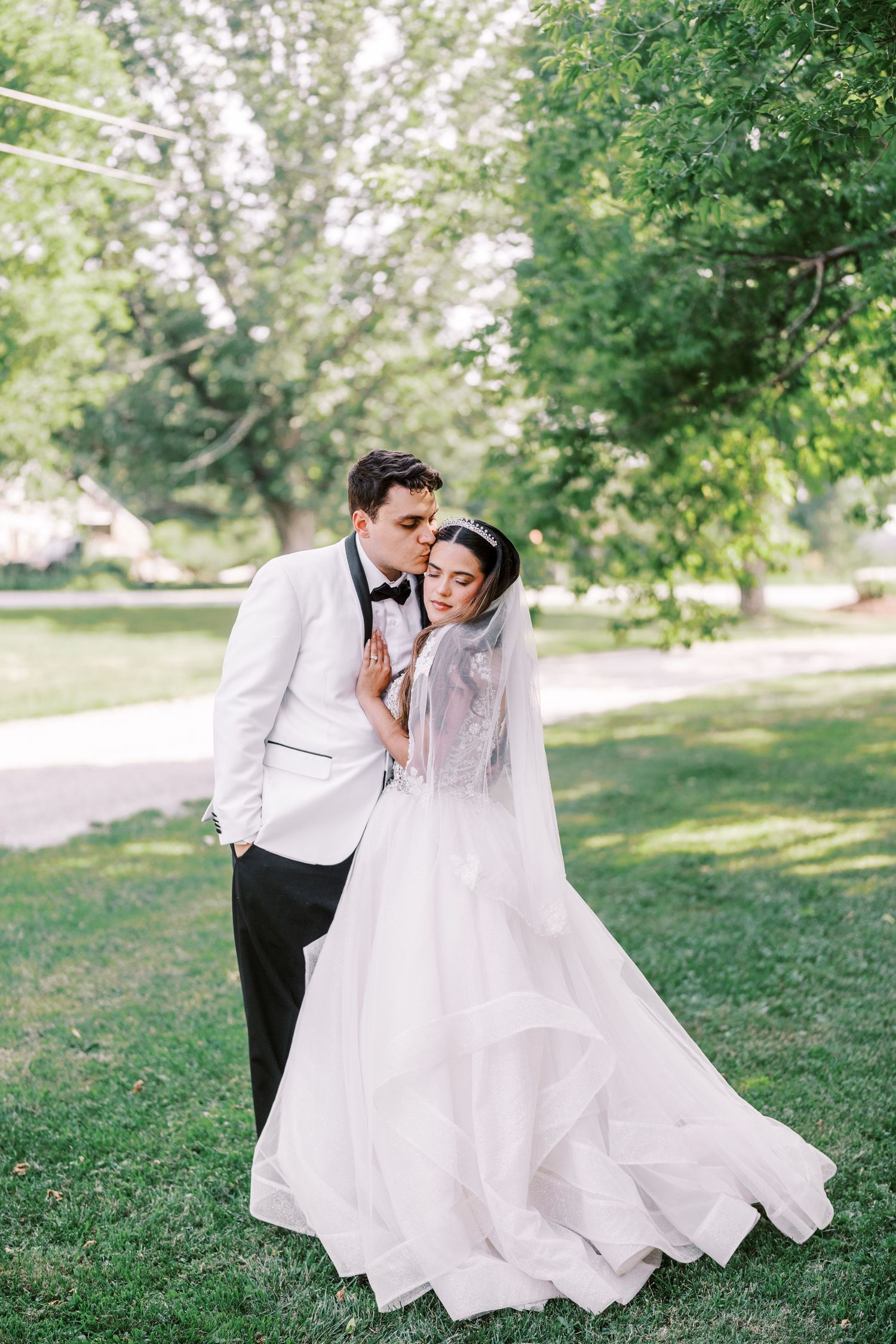 A romantic outdoor wedding portrait of a couple in white formal attire with a flowing bridal veil in a lush garden setting.