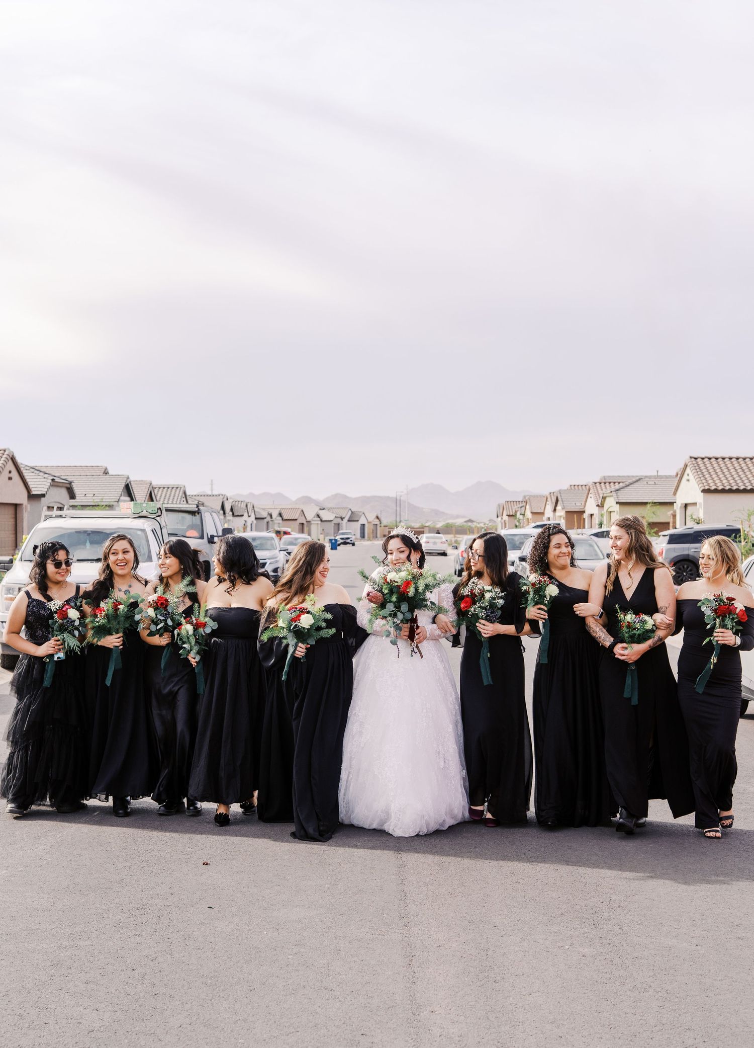 Wedding party in black bridesmaid dresses and white bridal gown pose together on suburban street with houses in background.