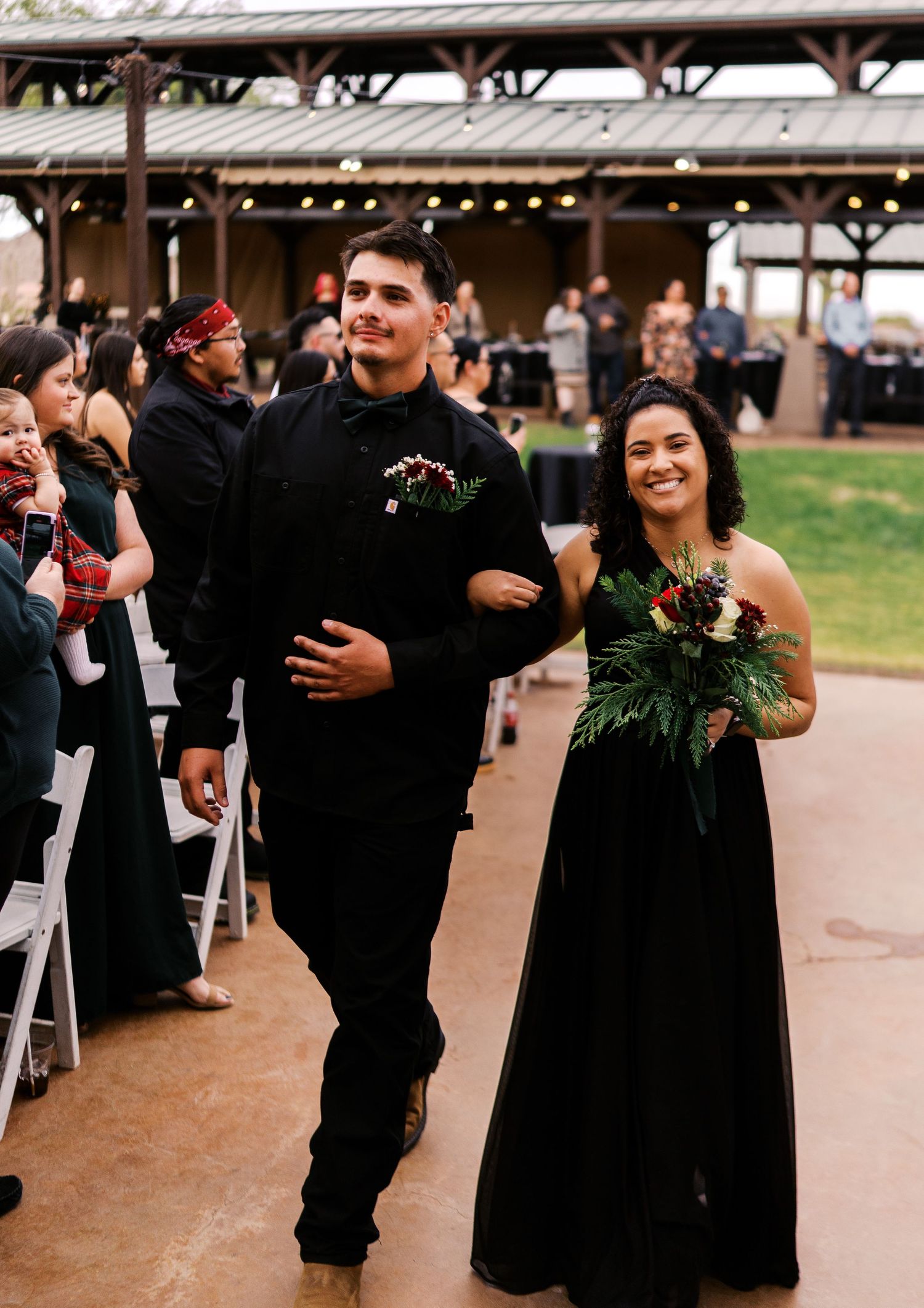 A couple in formal attire walking down an outdoor aisle at a wedding celebration with guests seated on either side.