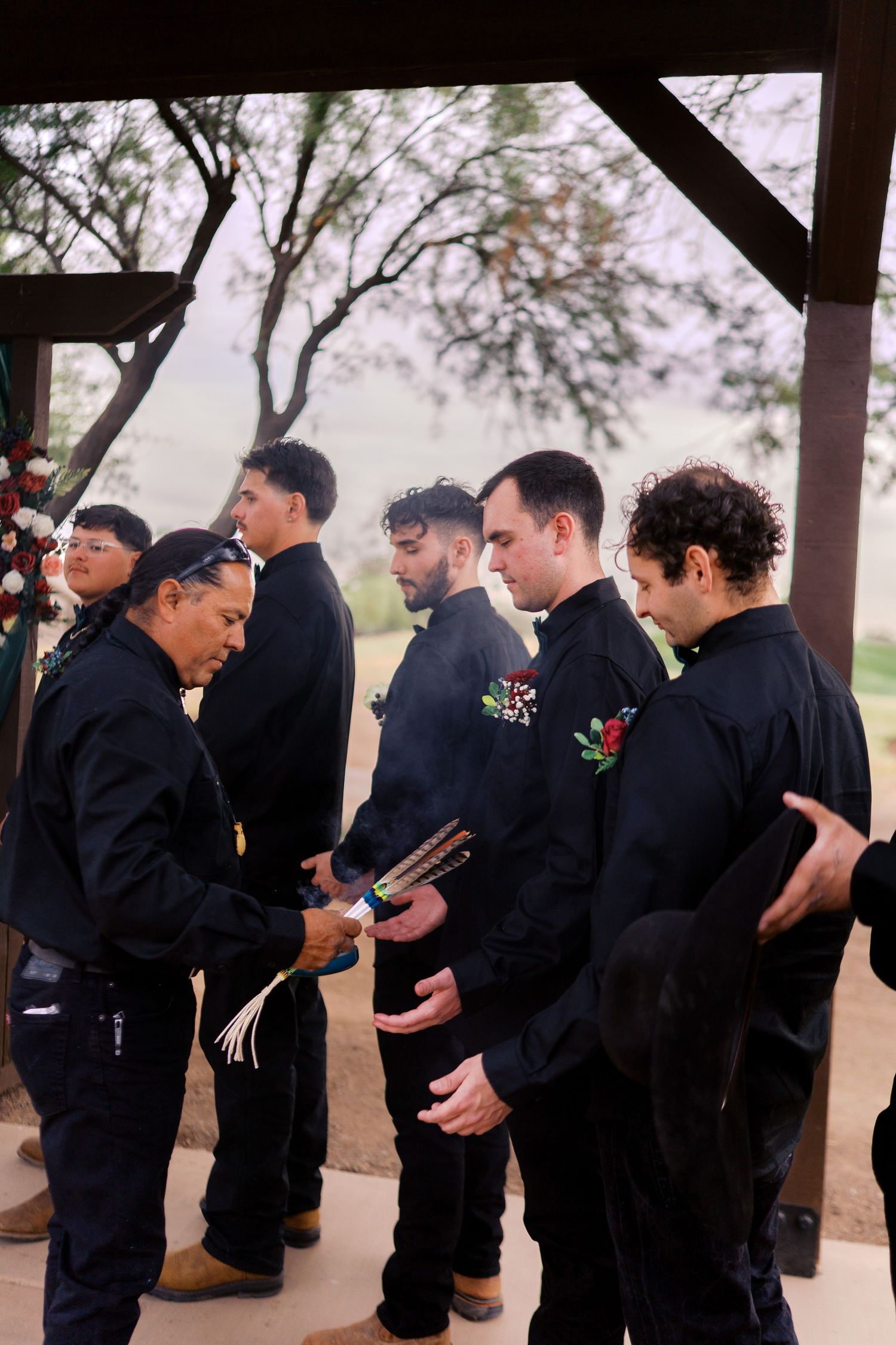 A group of groomsmen in black suits with boutonnieres stand together at an outdoor ceremony.