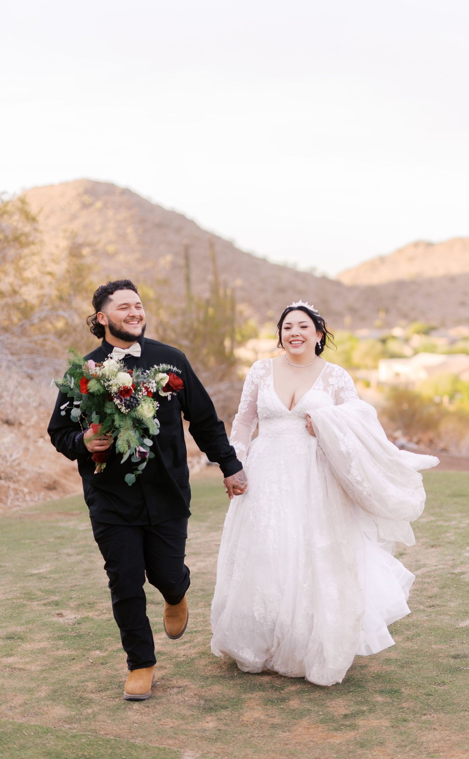 A happy couple runs together across desert terrain, with the bride in white dress and groom holding a floral bouquet.