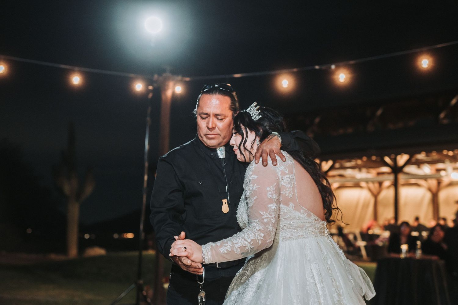 A couple shares their first dance under twinkling string lights at an outdoor evening wedding reception.