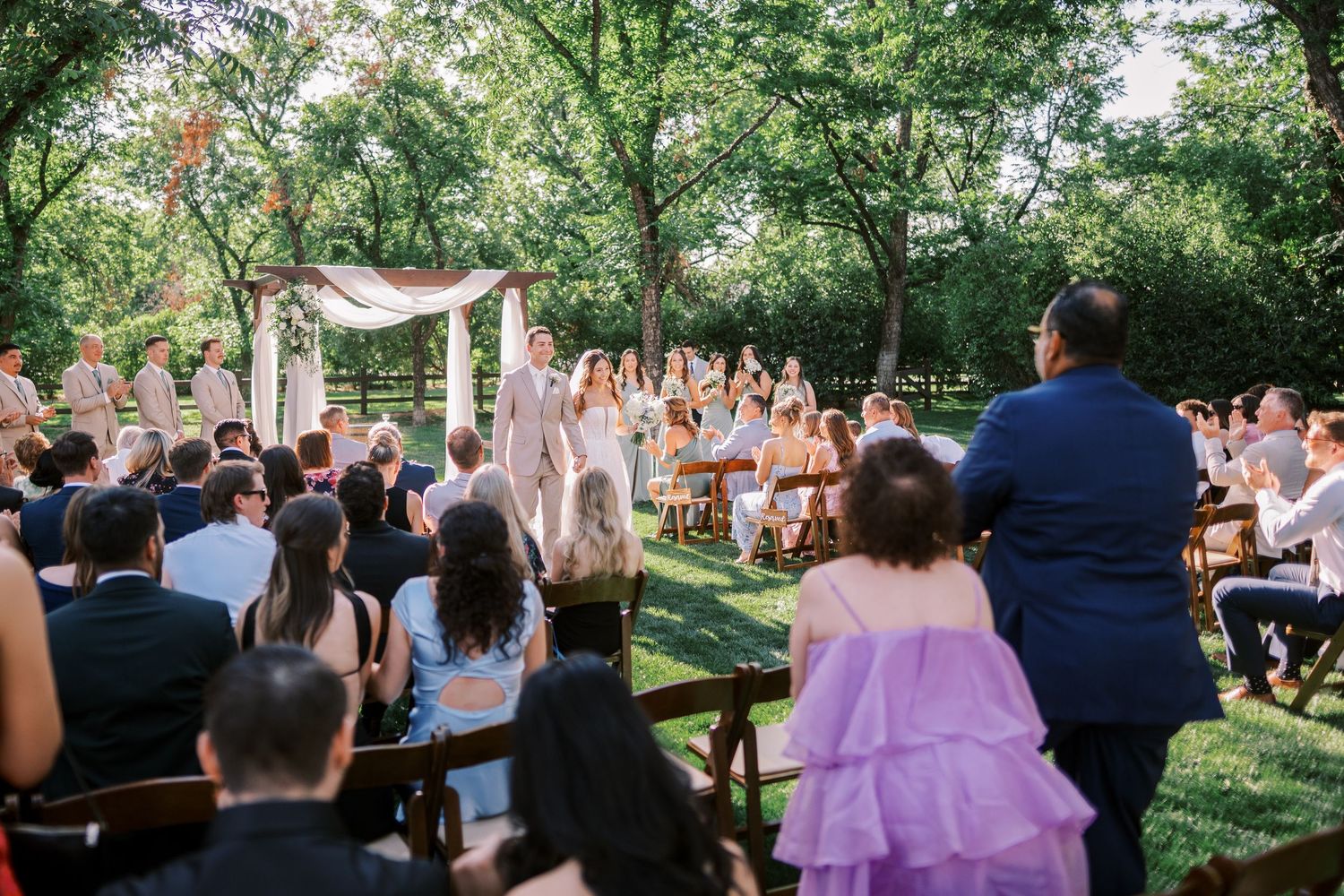 An outdoor wedding ceremony takes place in a lush green garden setting with guests seated on both sides of a decorated aisle.