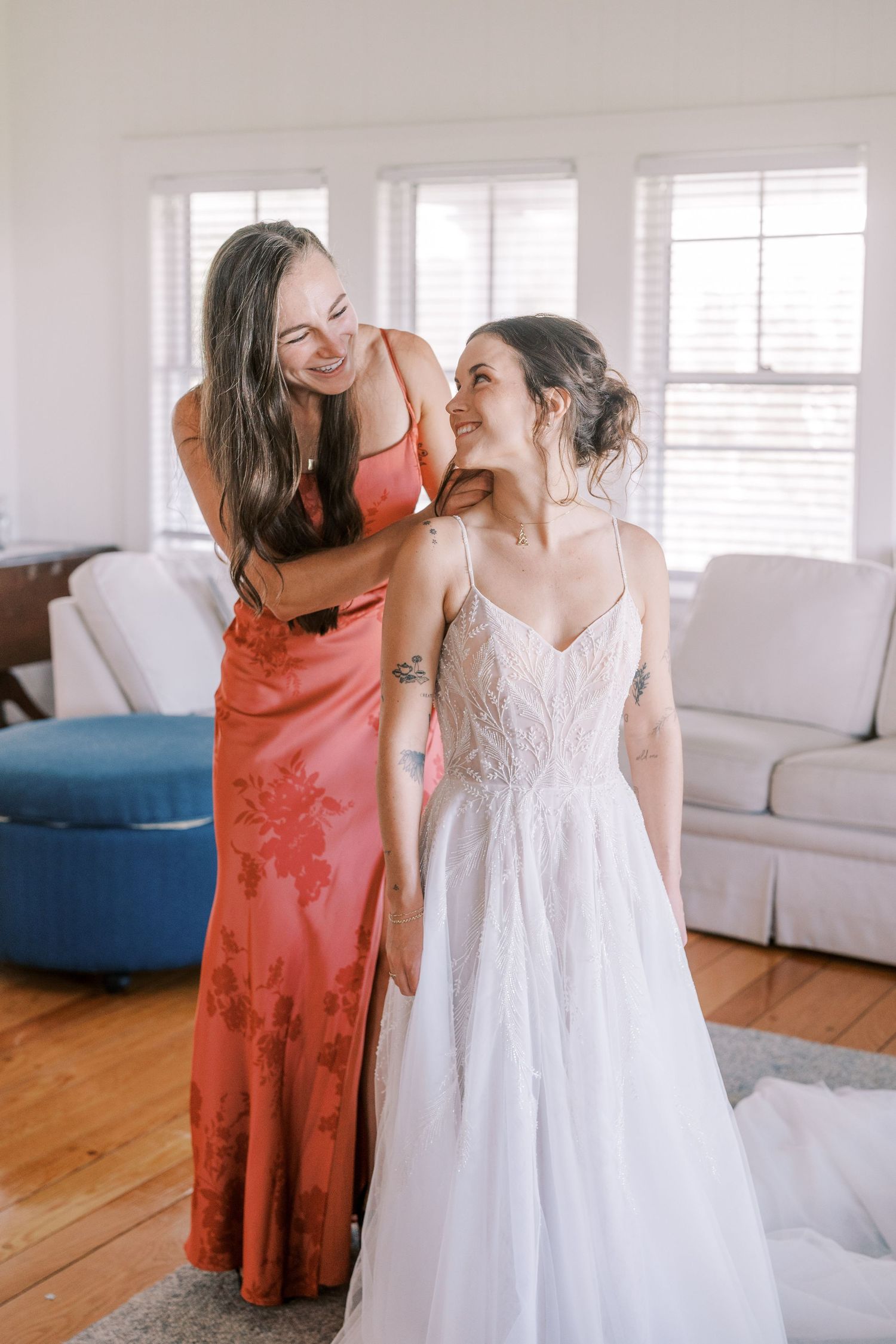Candid pre-wedding moment shared between women in formal dresses in sunlit room.