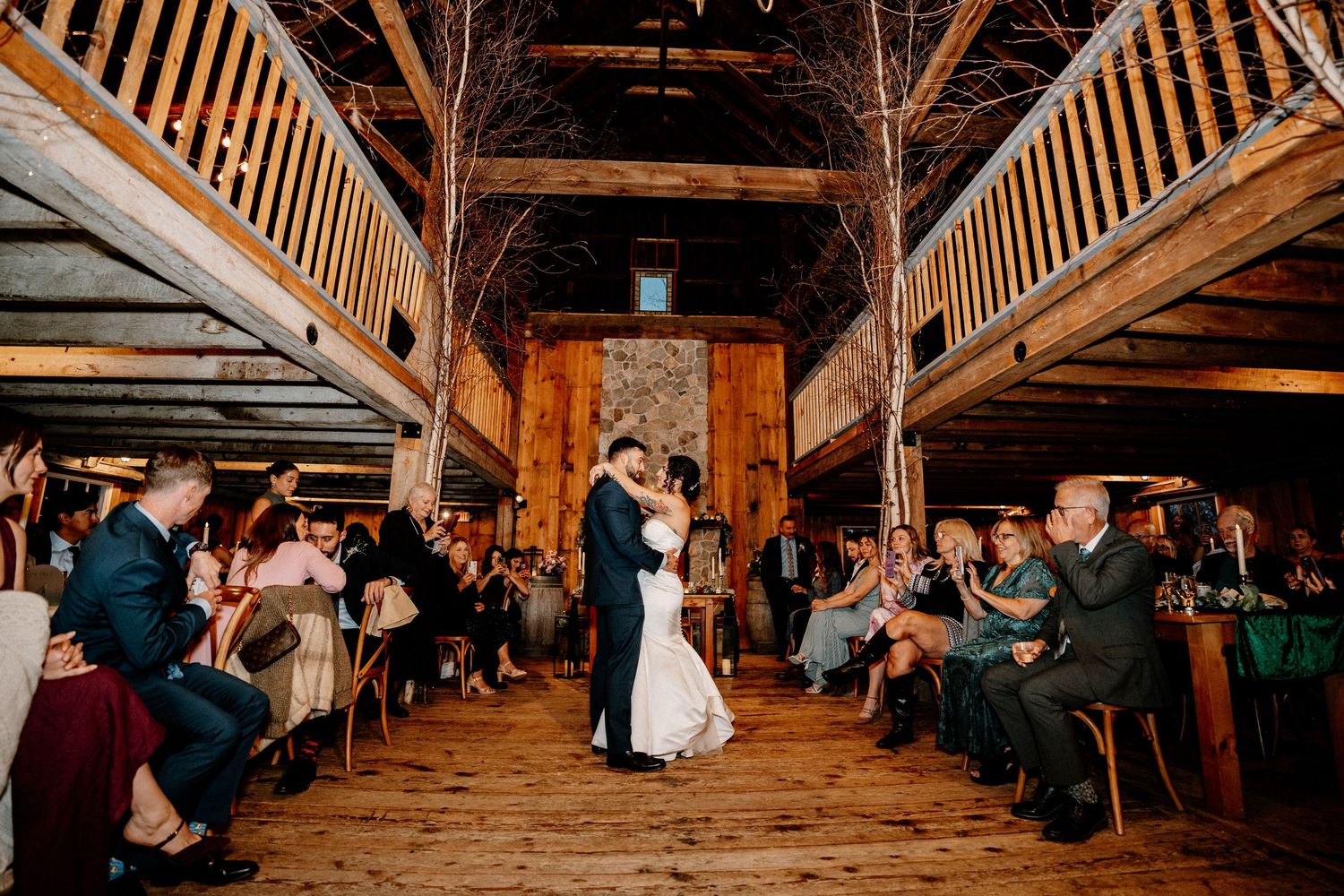 Wedding guests dance and celebrate inside a rustic wooden barn venue with exposed beams and warm lighting.