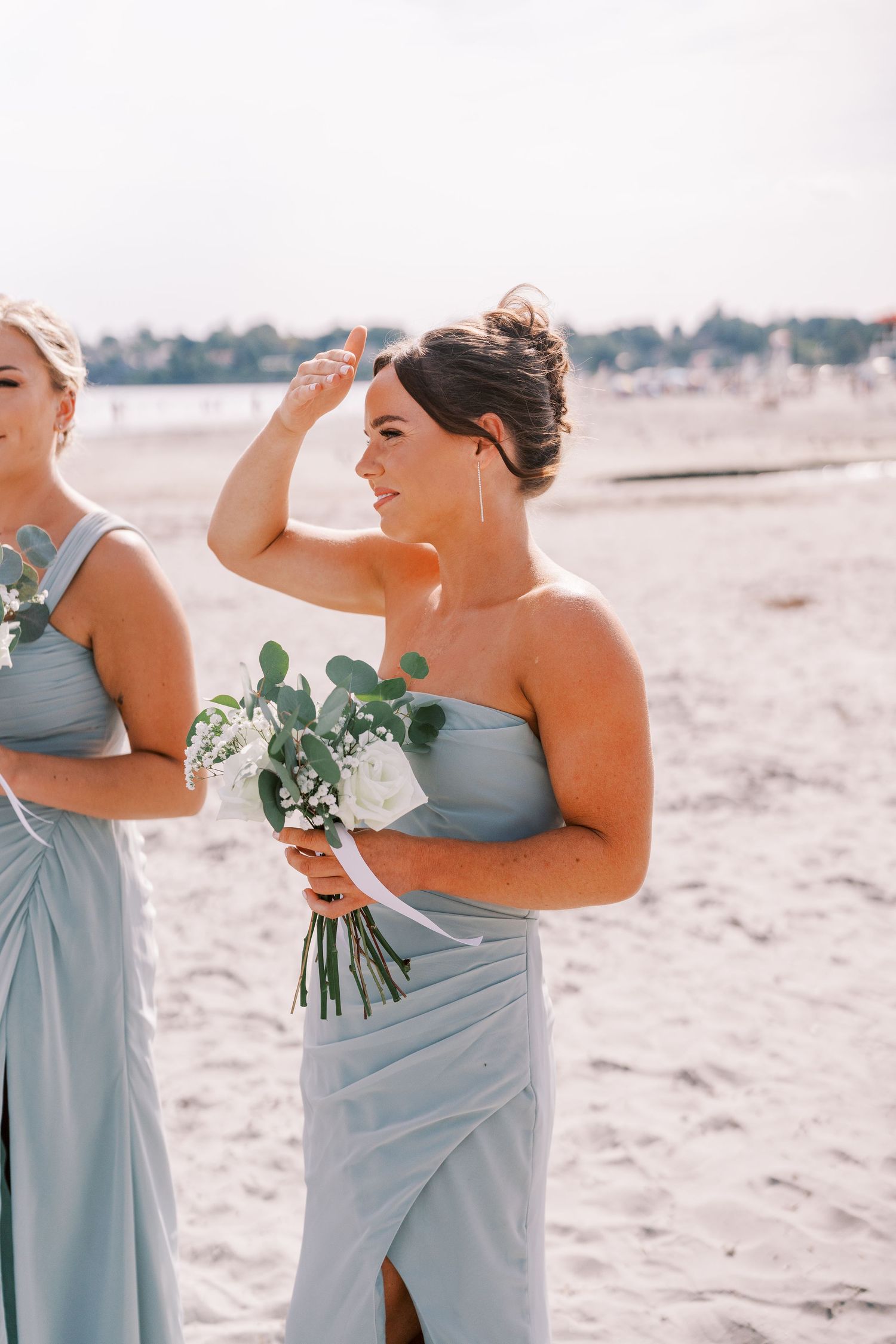 Bridesmaids in light blue dresses laugh together while holding eucalyptus bouquets on a sunny beach.