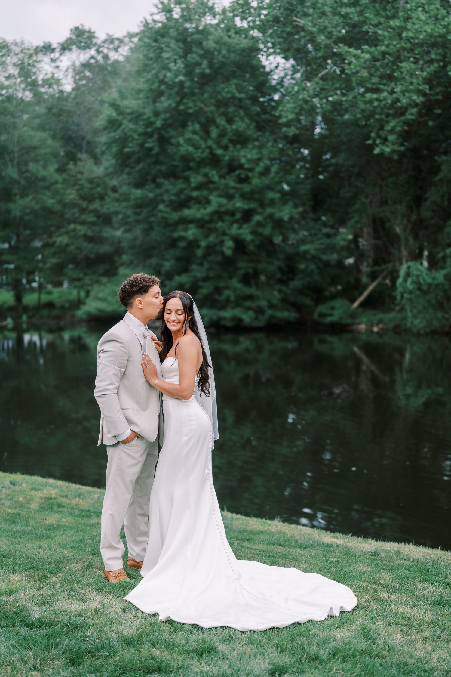 A wedding couple embraces on a grassy lawn beside a serene pond surrounded by trees.