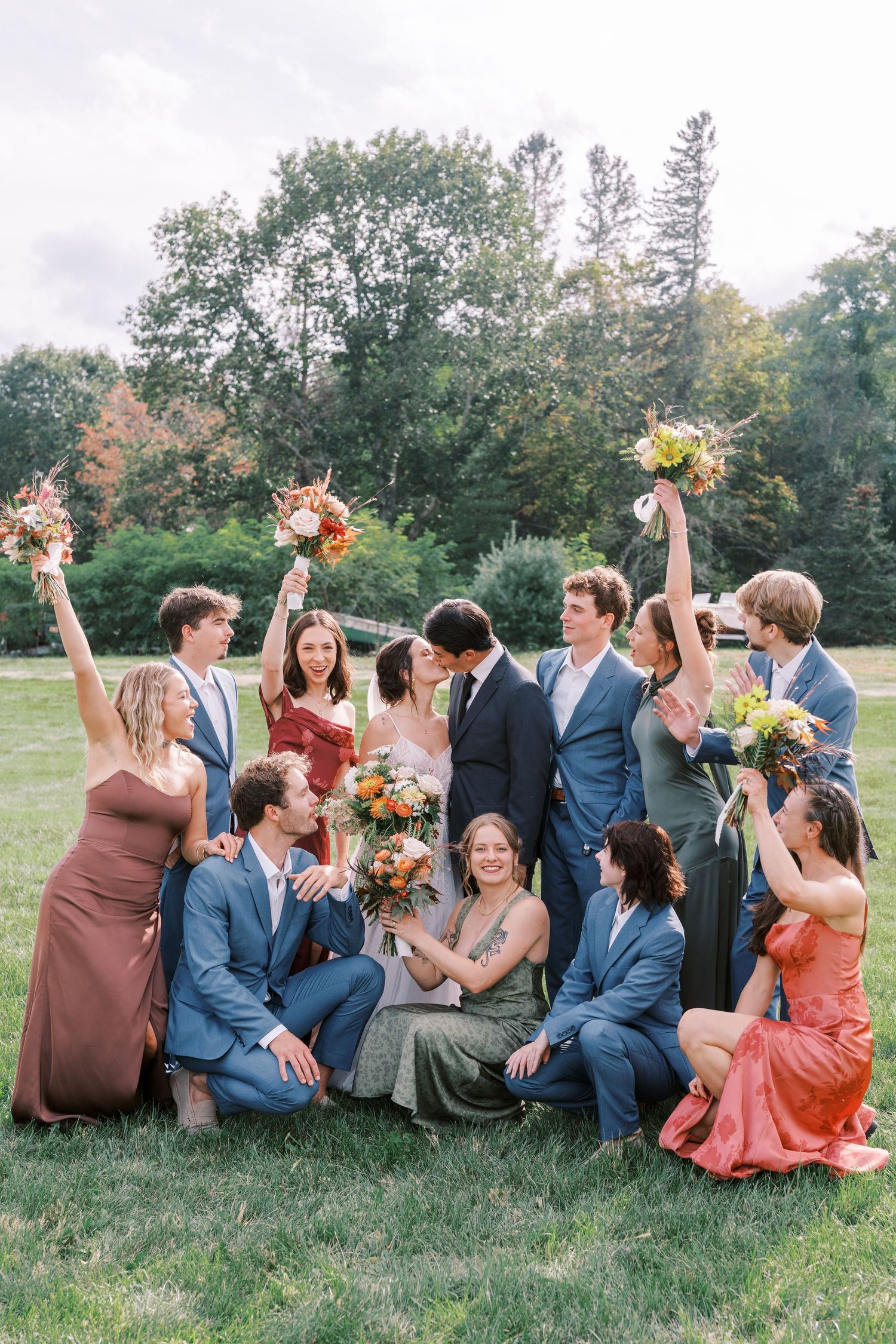 Wedding party in blue suits and coral dresses poses playfully on grassy hillside.