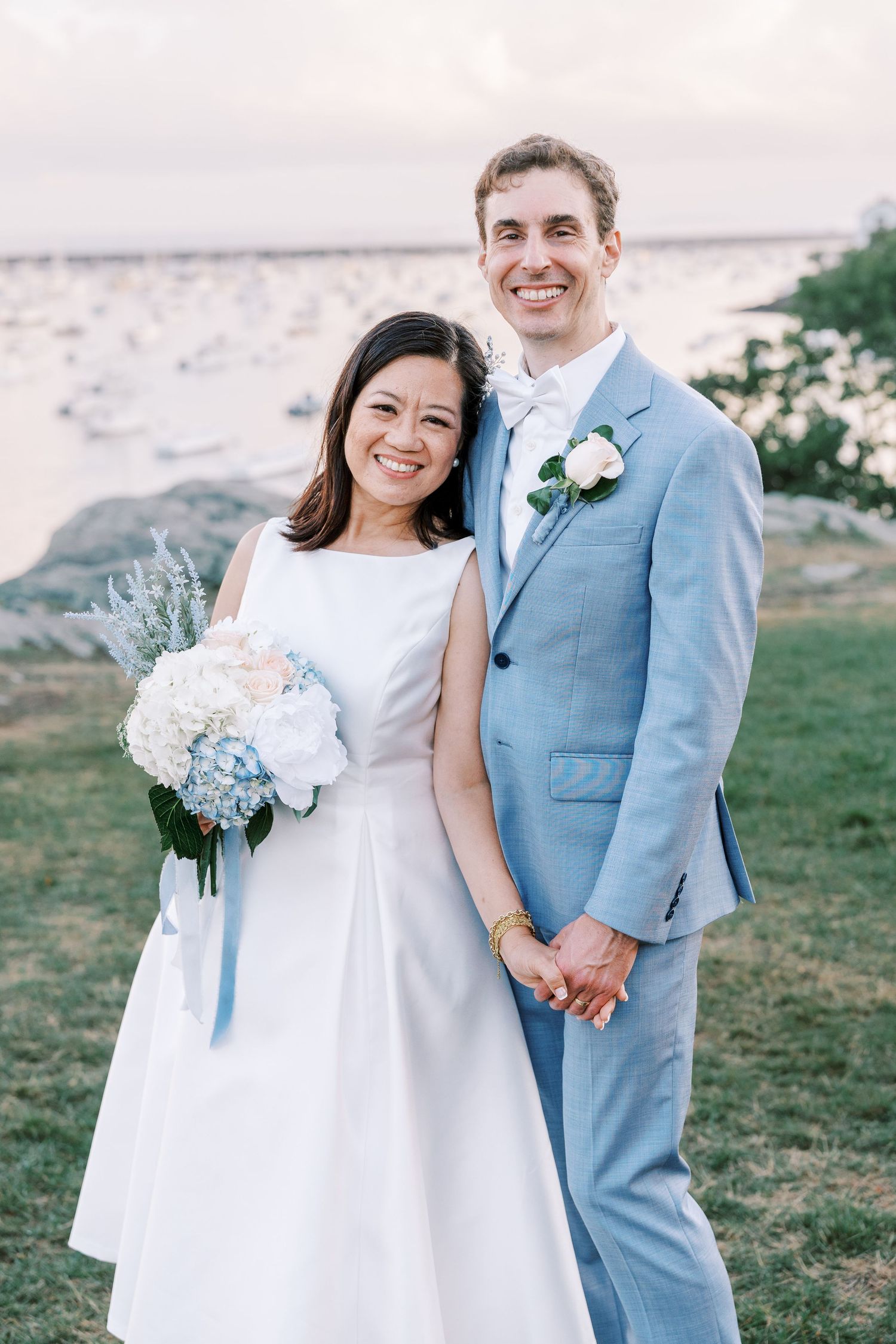 Newlywed couple in powder blue suit and white dress pose together outdoors for wedding photos against coastal scenery.