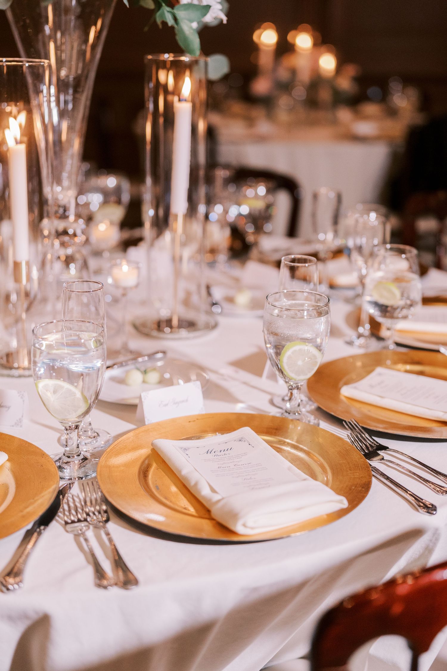 Elegant wedding reception table setting with gold charger plates, white linens, and crystal glassware illuminated by candlelight.