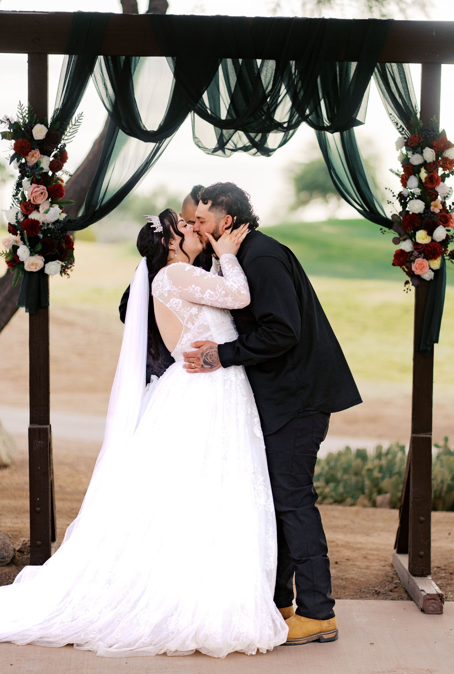 A wedding ceremony under a wooden arch with dark green draping and floral arrangements outdoors.