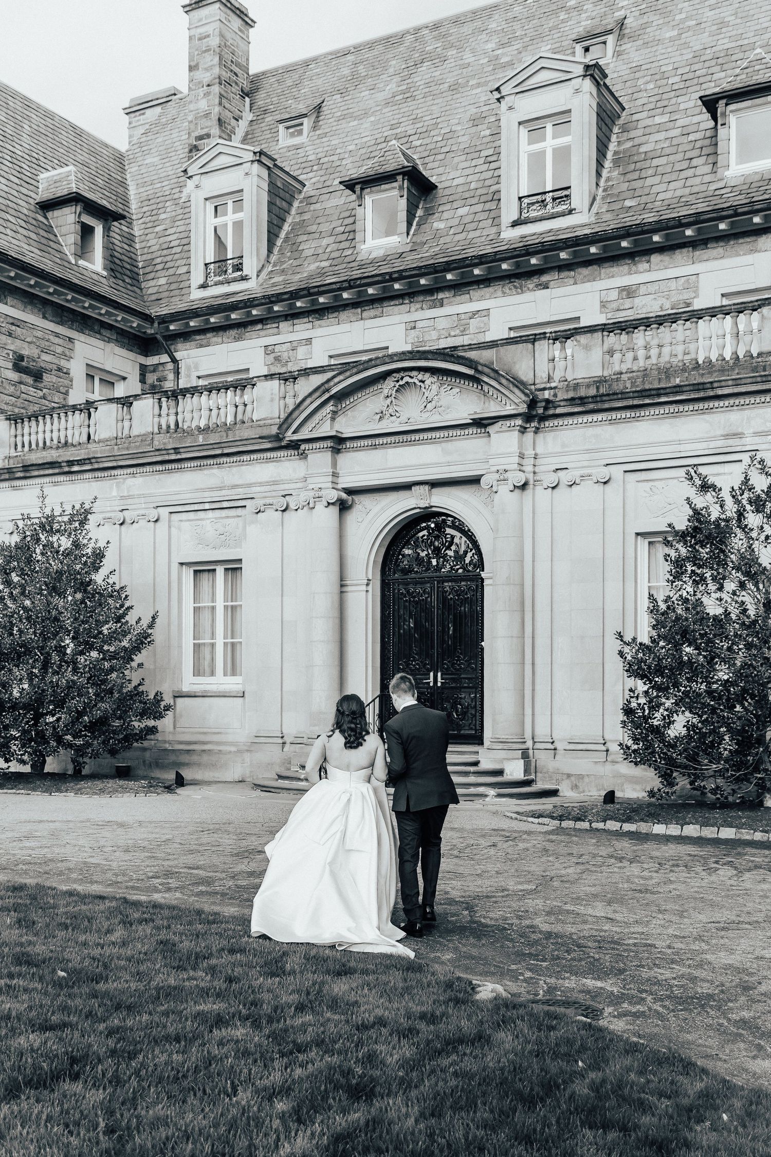 Black and white photo of a couple walking towards grand mansion entrance on their wedding day.