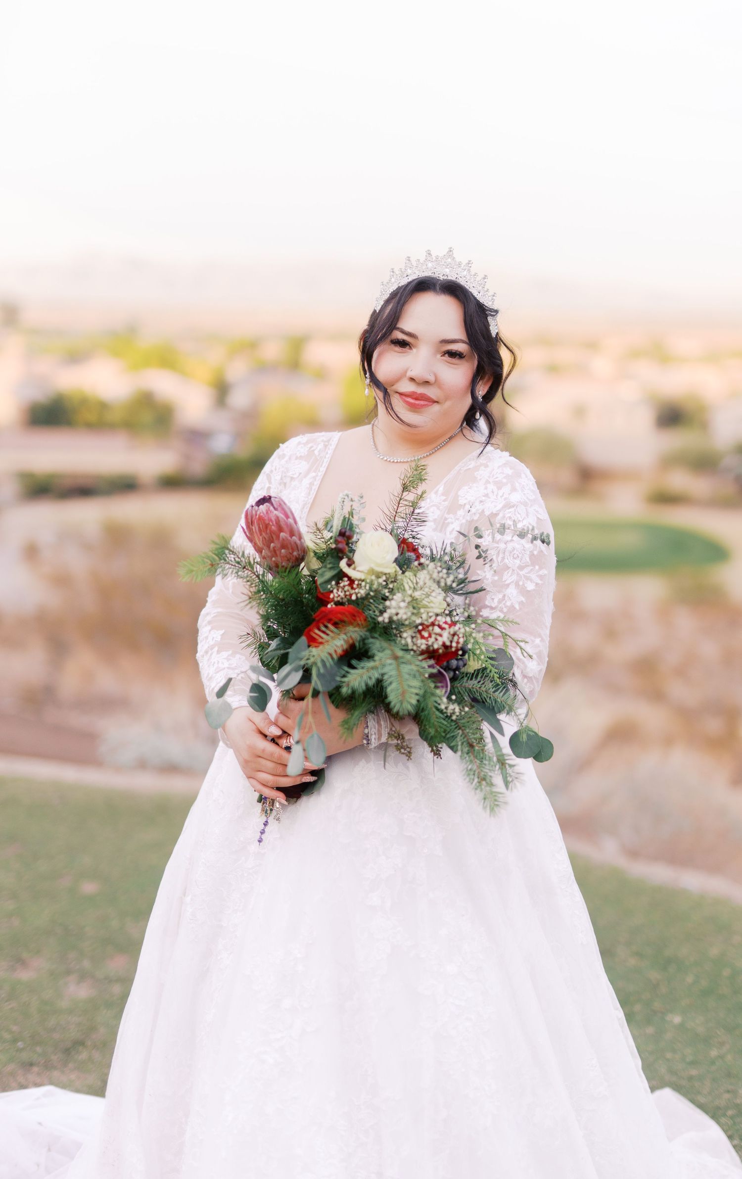 A bride in a white lace gown holds a vibrant bouquet featuring protea flowers and greenery against a sunset backdrop.