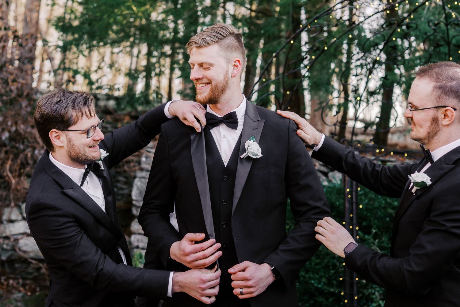 Three groomsmen laughing together while adjusting their formal attire against a backdrop of evergreen trees.
