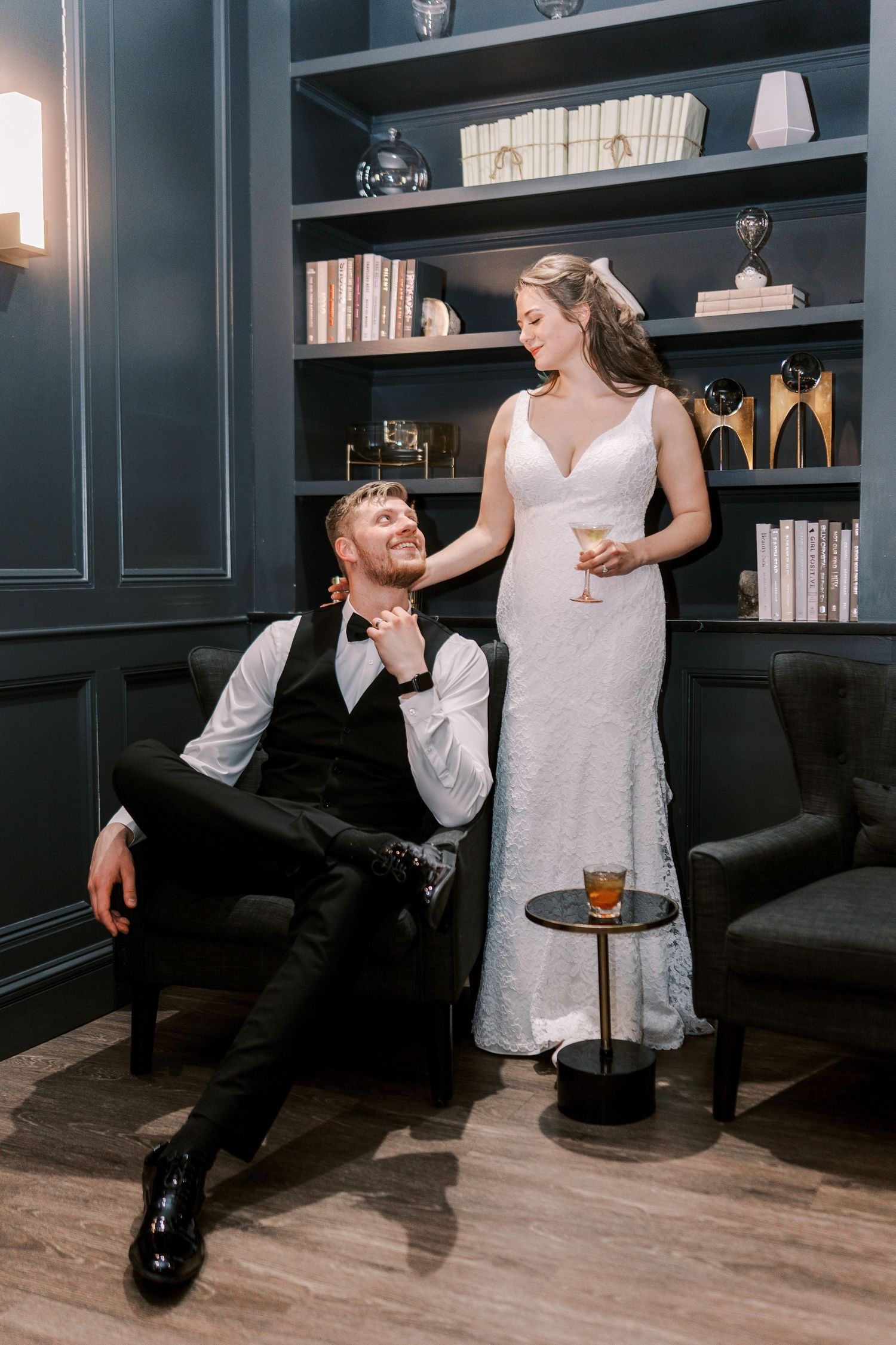 A couple in wedding attire shares a playful moment in an elegant library setting with dark built-in bookshelves.