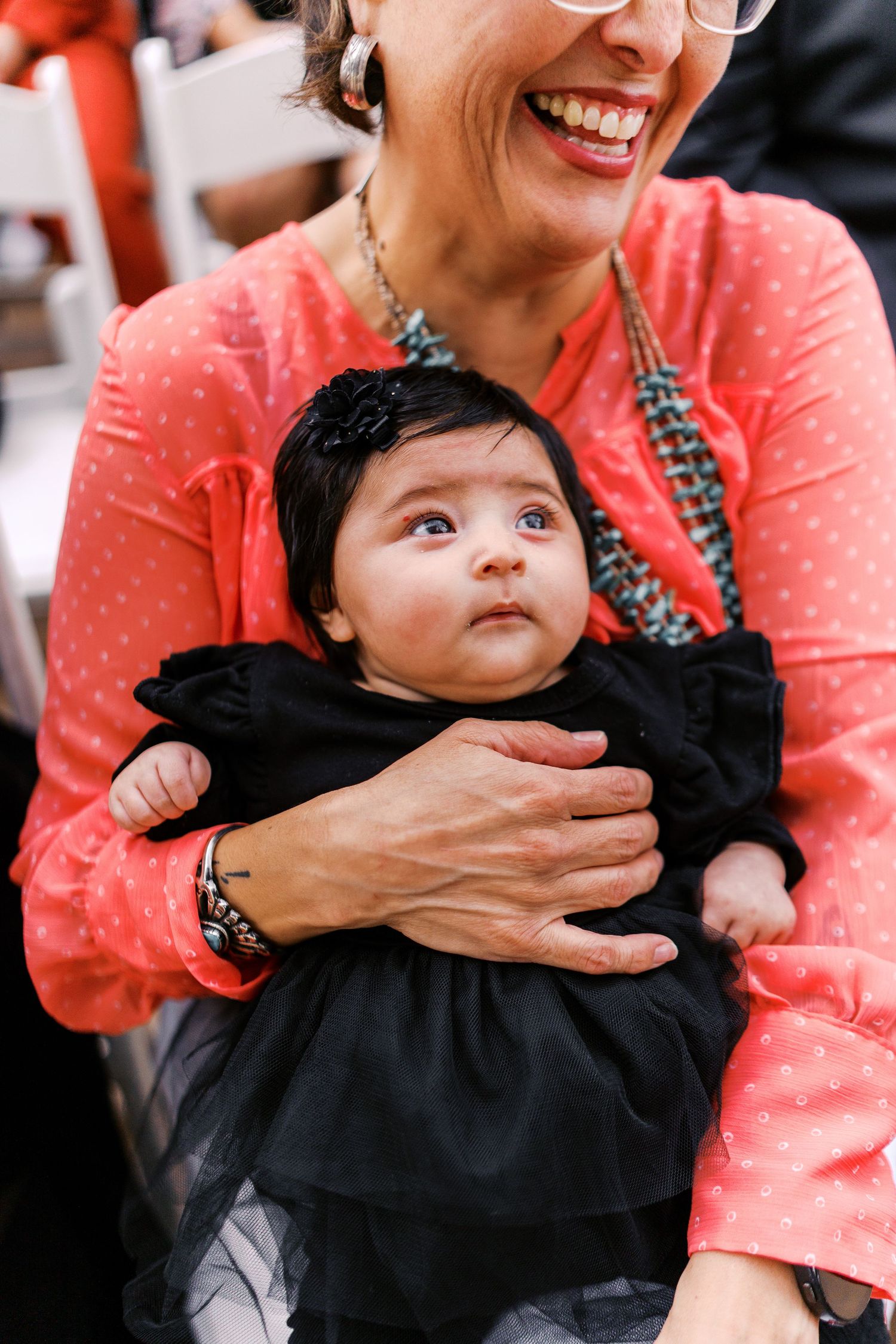 An adorable baby in a black outfit looks up curiously while being held by someone wearing a coral polka dot top.