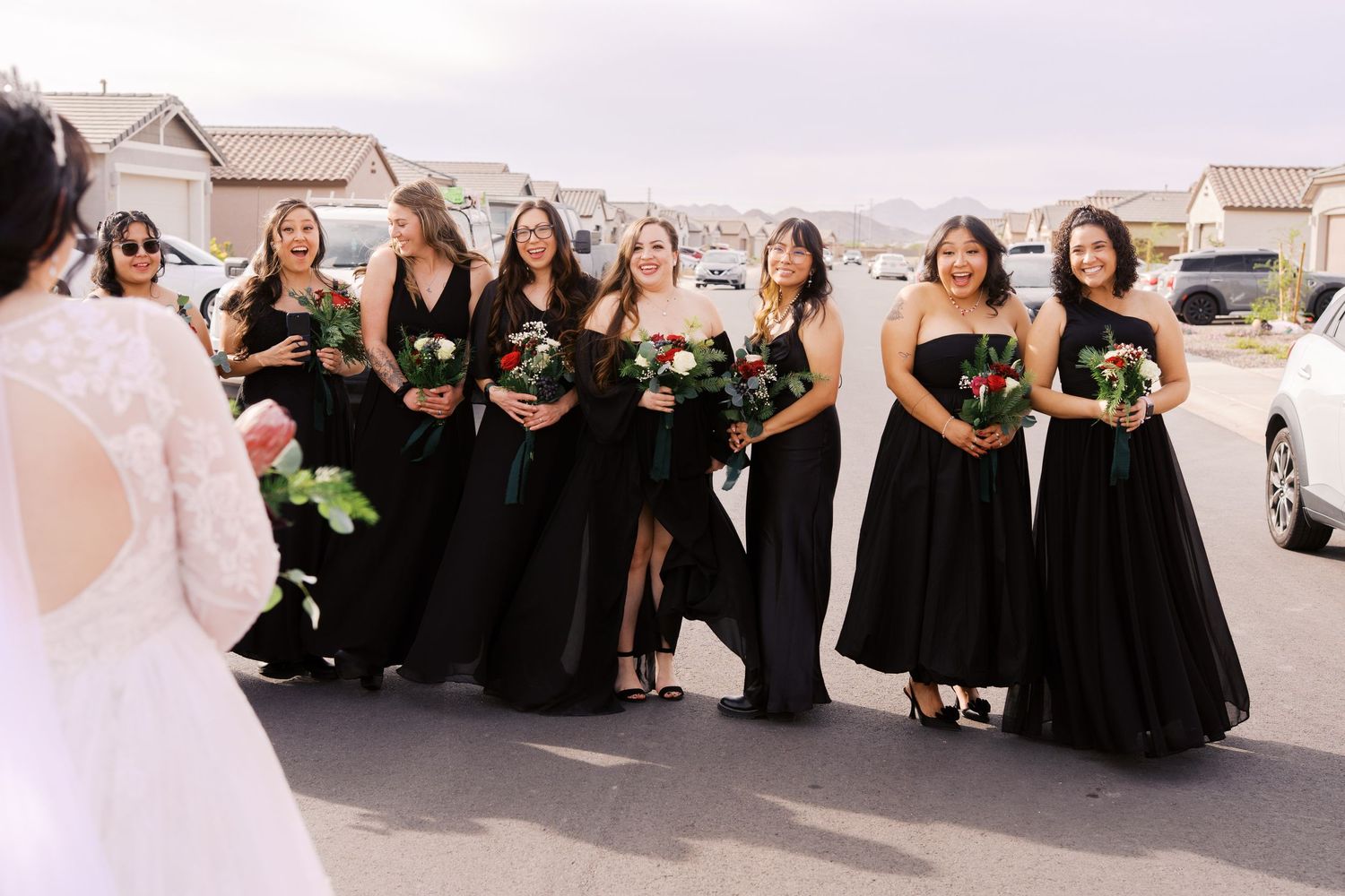 A bridal party wearing elegant black floor-length gowns poses outside in a row while holding floral bouquets.