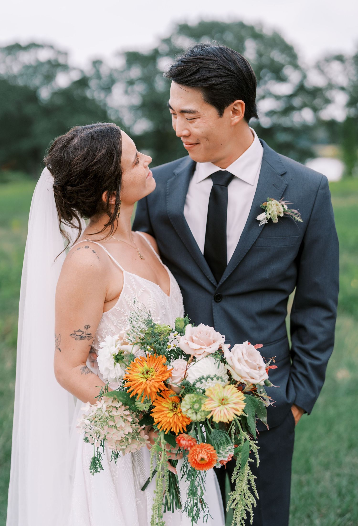 Romantic wedding couple poses outdoors with vibrant orange and yellow floral bouquet in a garden setting.