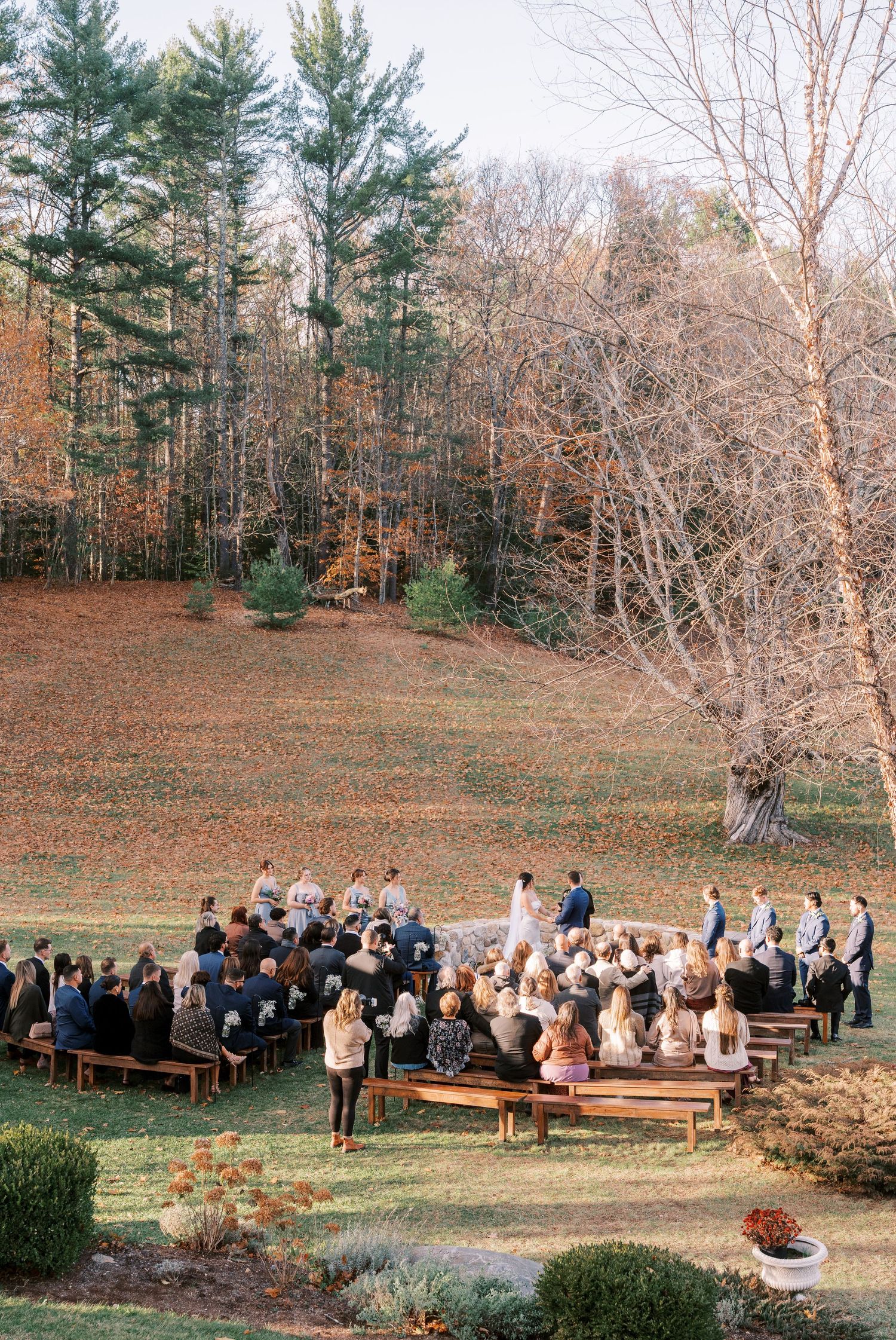 Outdoor wedding ceremony in a wooded clearing with guests seated on rustic wooden benches during autumn.