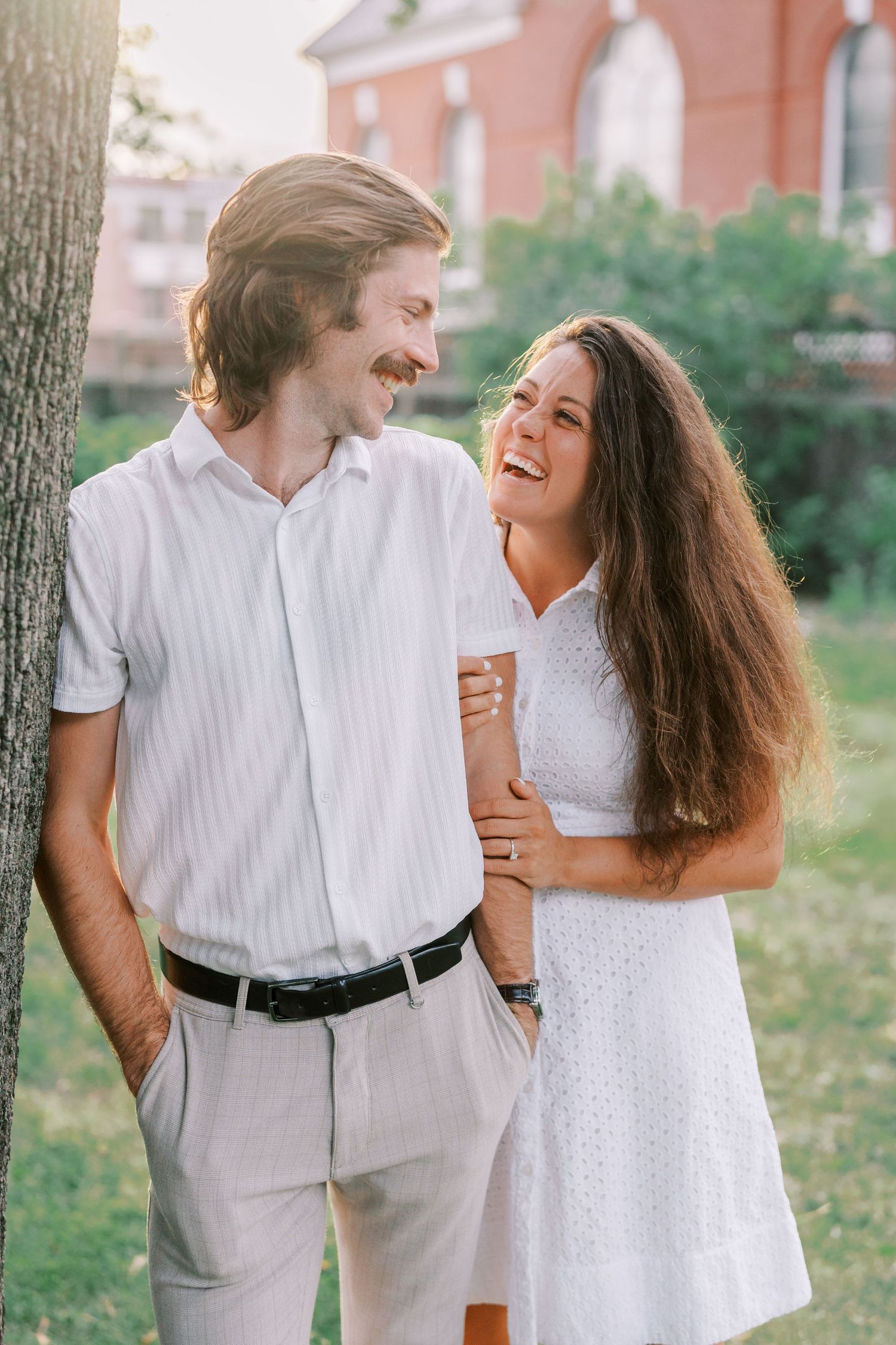 A couple in white outfits laughs together while embracing in a sunny outdoor setting.