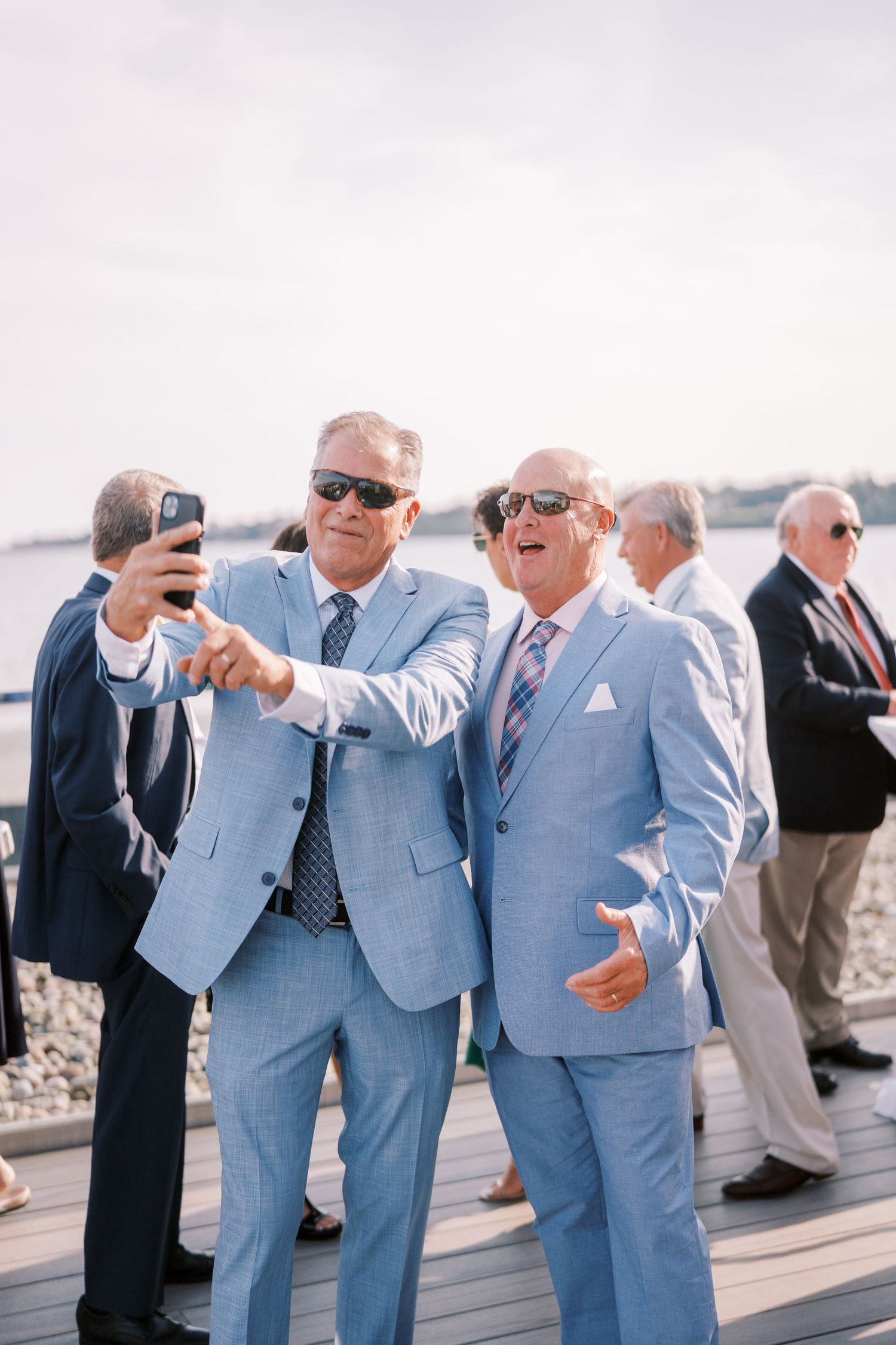 Group of men in light blue suits laughing and taking photos together on a boardwalk by the ocean.