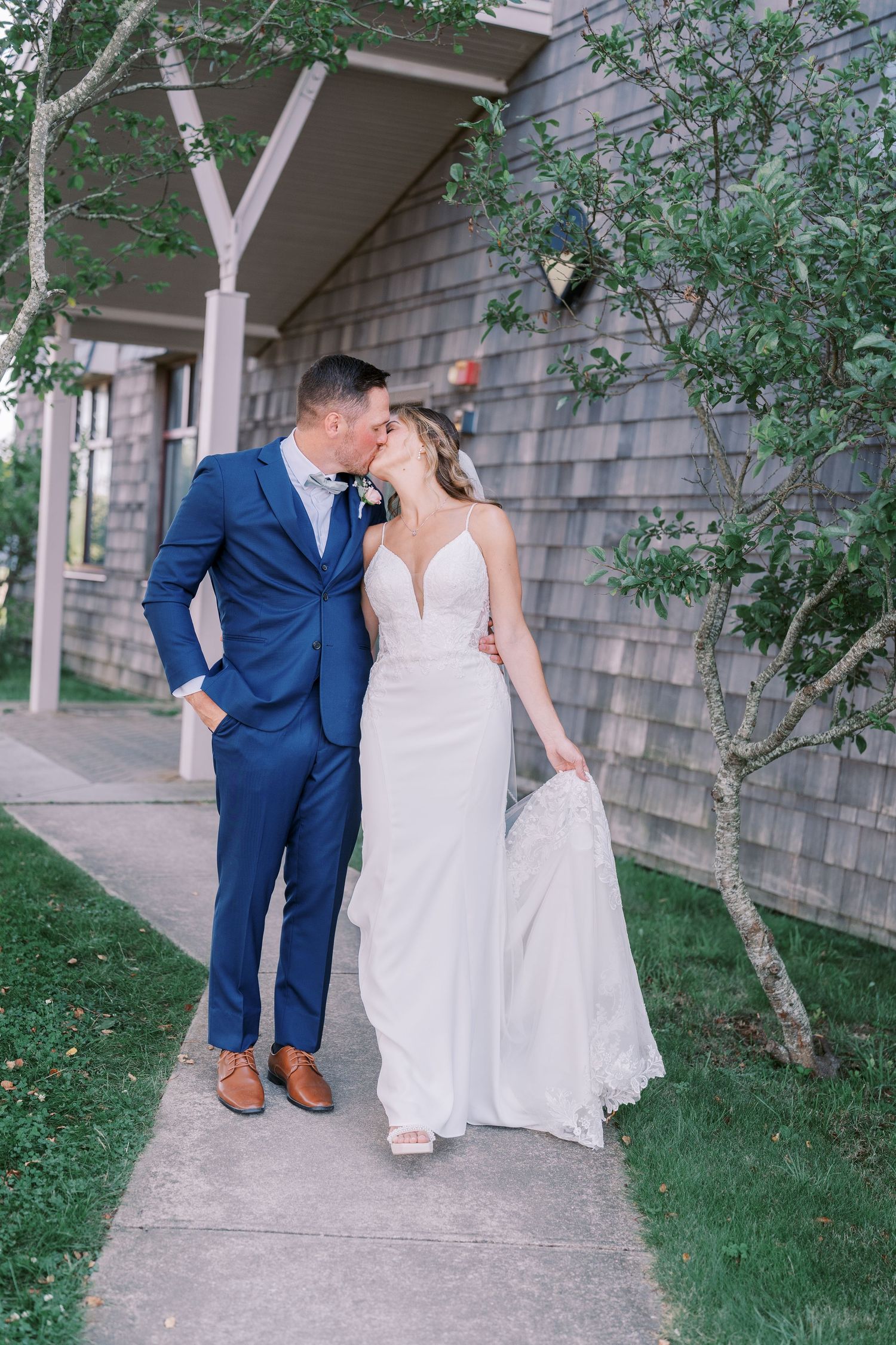 A couple in wedding attire shares an intimate moment on a walkway surrounded by greenery and rustic architecture.