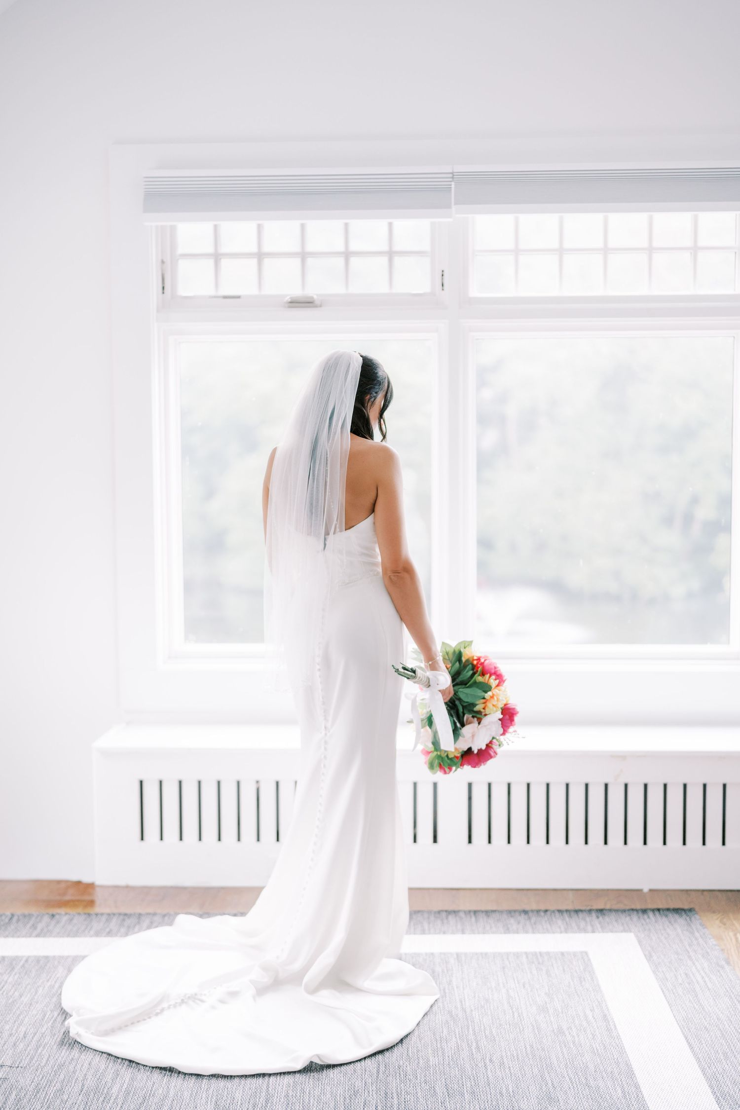 A wedding dress and veil silhouetted against a bright window while holding a colorful bouquet.