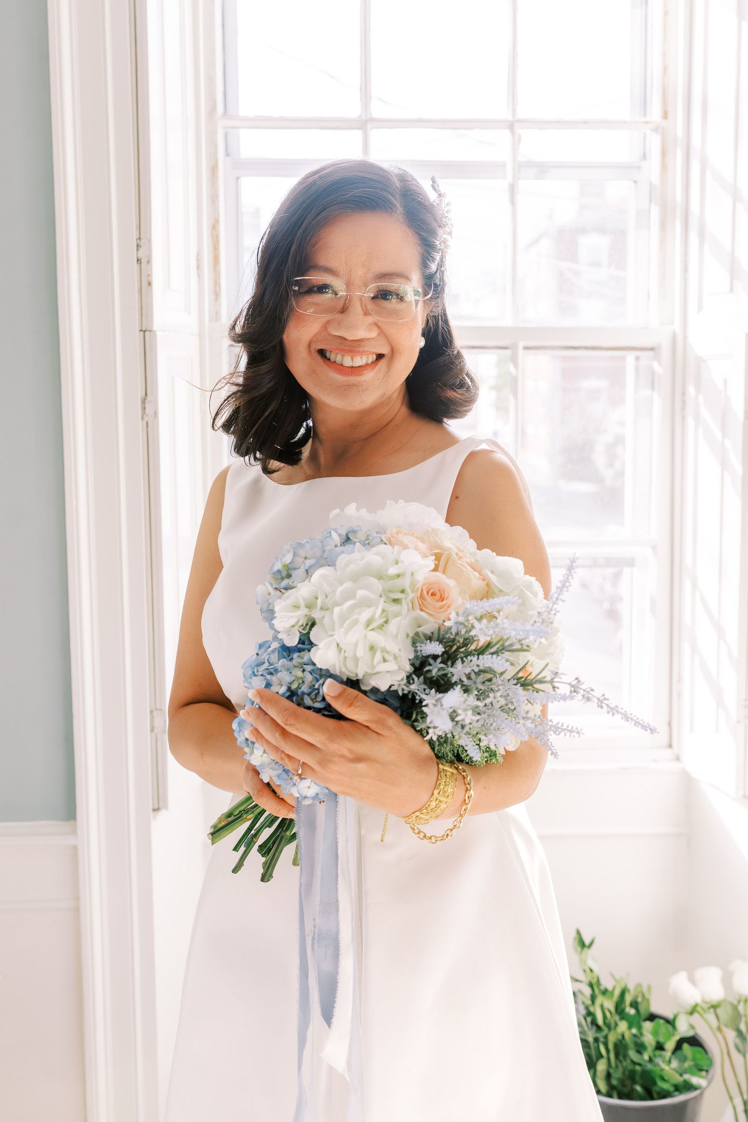 Bride in white dress holding elegant bouquet of white and blue flowers by bright windows.