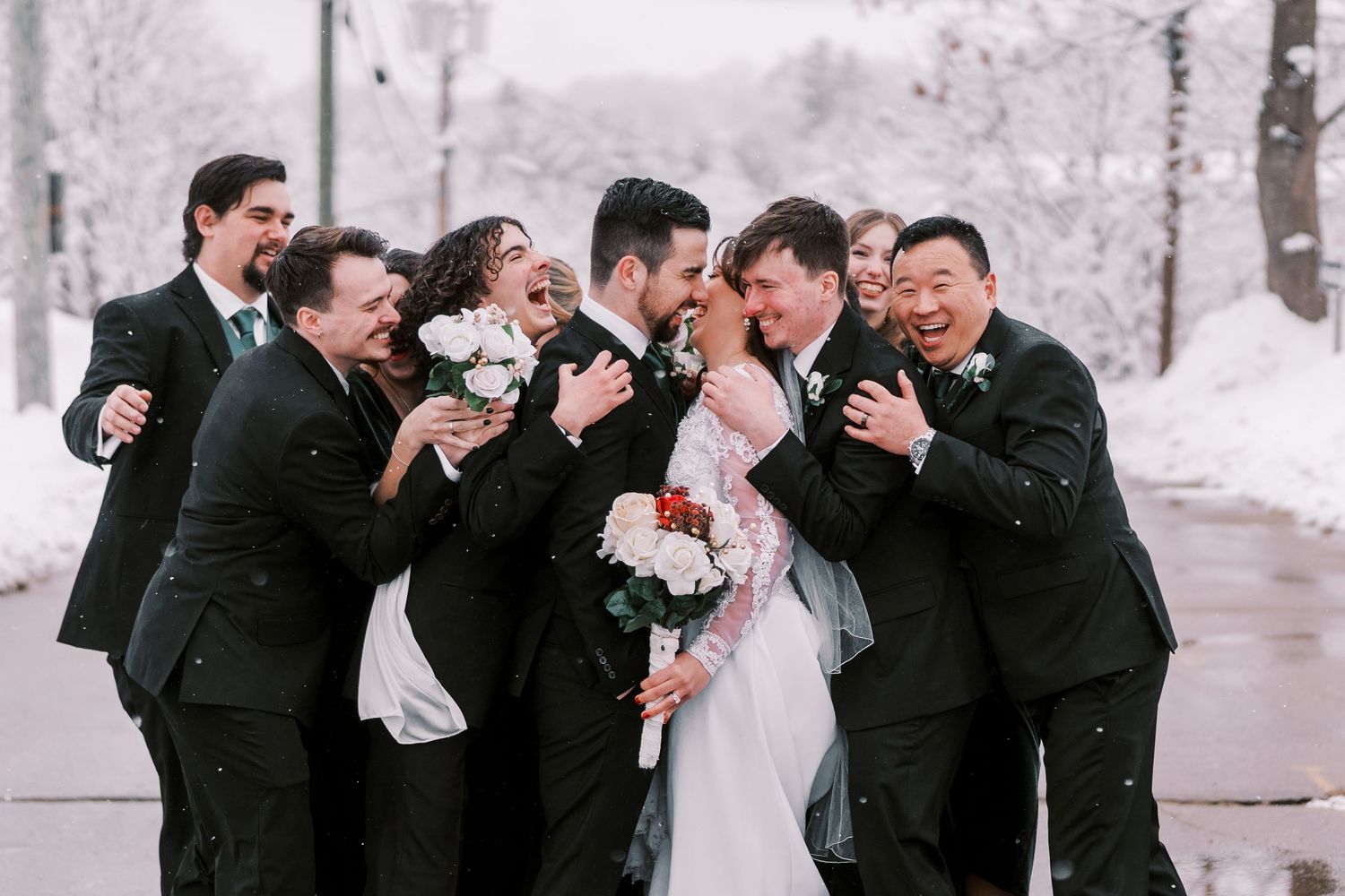 A wedding party in black suits laughs together during a snowy winter photo session outdoors.