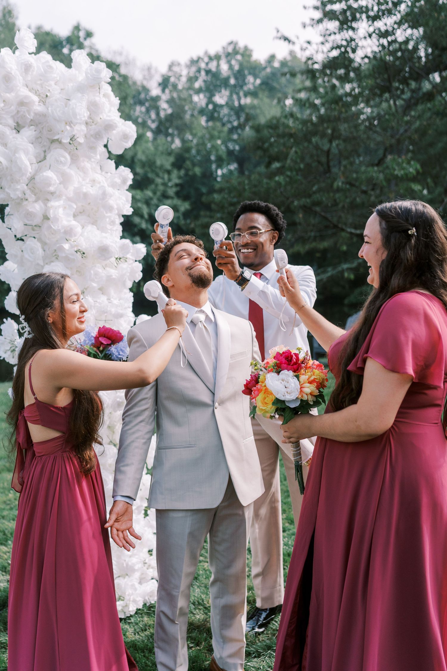 Wedding party in burgundy dresses and grey suit laugh while standing next to white balloon arch during outdoor celebration.