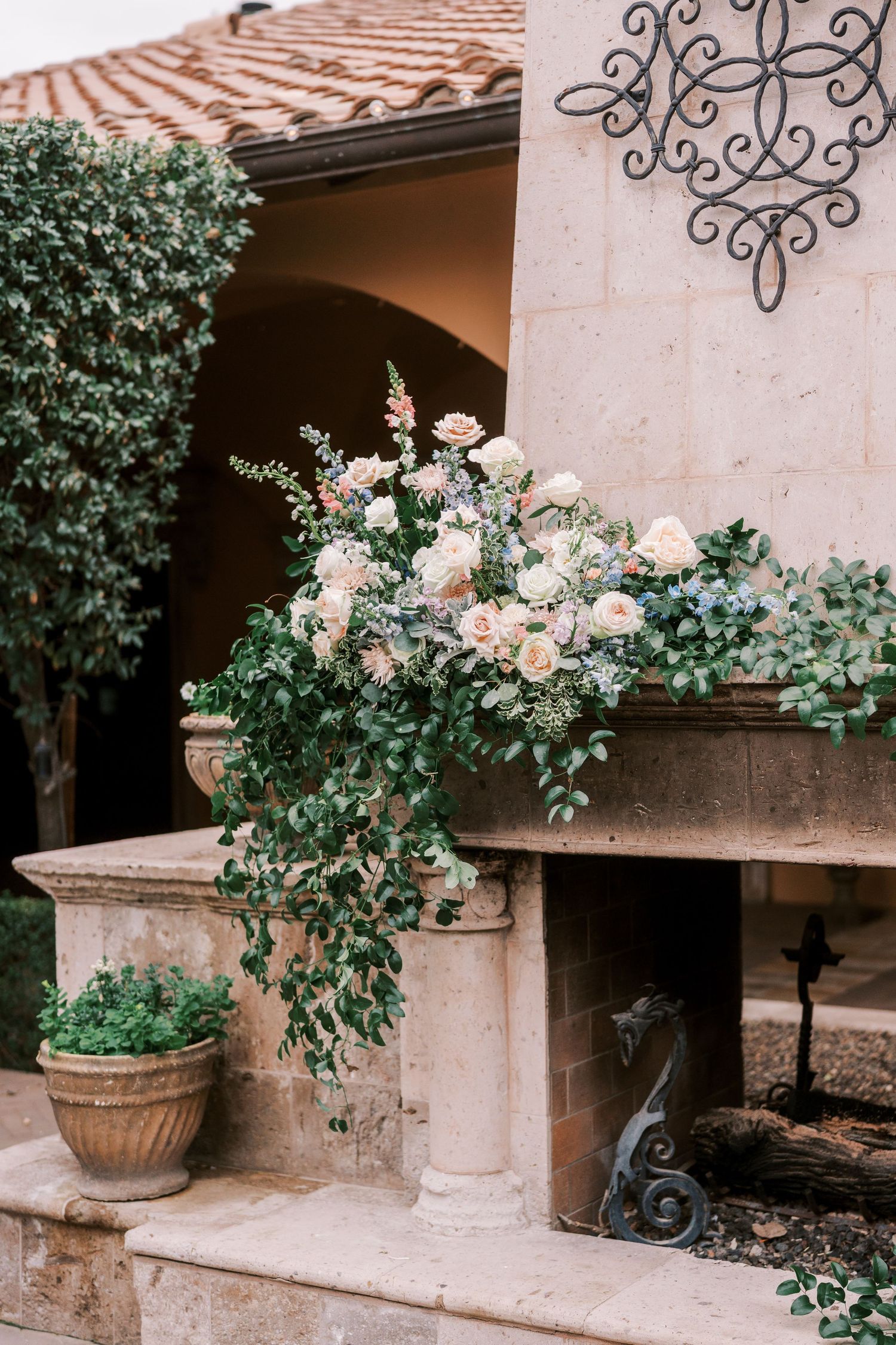 Elegant stone villa patio with potted plants, ornate iron scrollwork, and terracotta roof tiles in Mediterranean style.