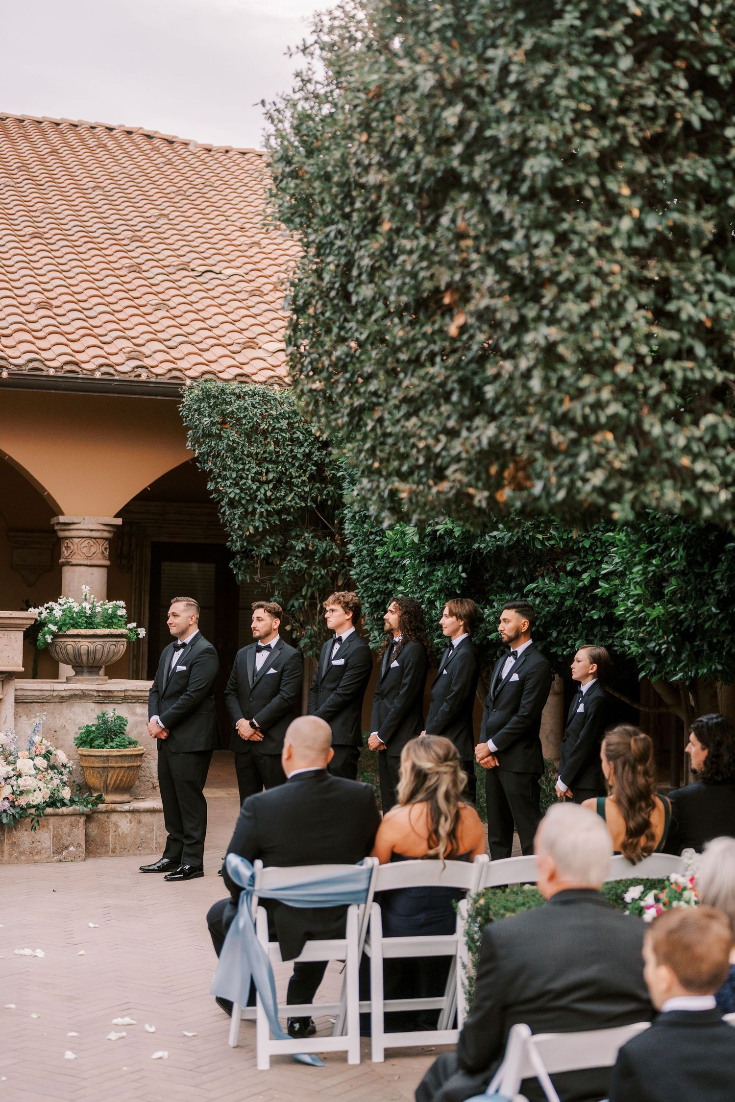 Outdoor wedding ceremony setup with white chairs and guests gathered under trees at a Spanish-style venue.