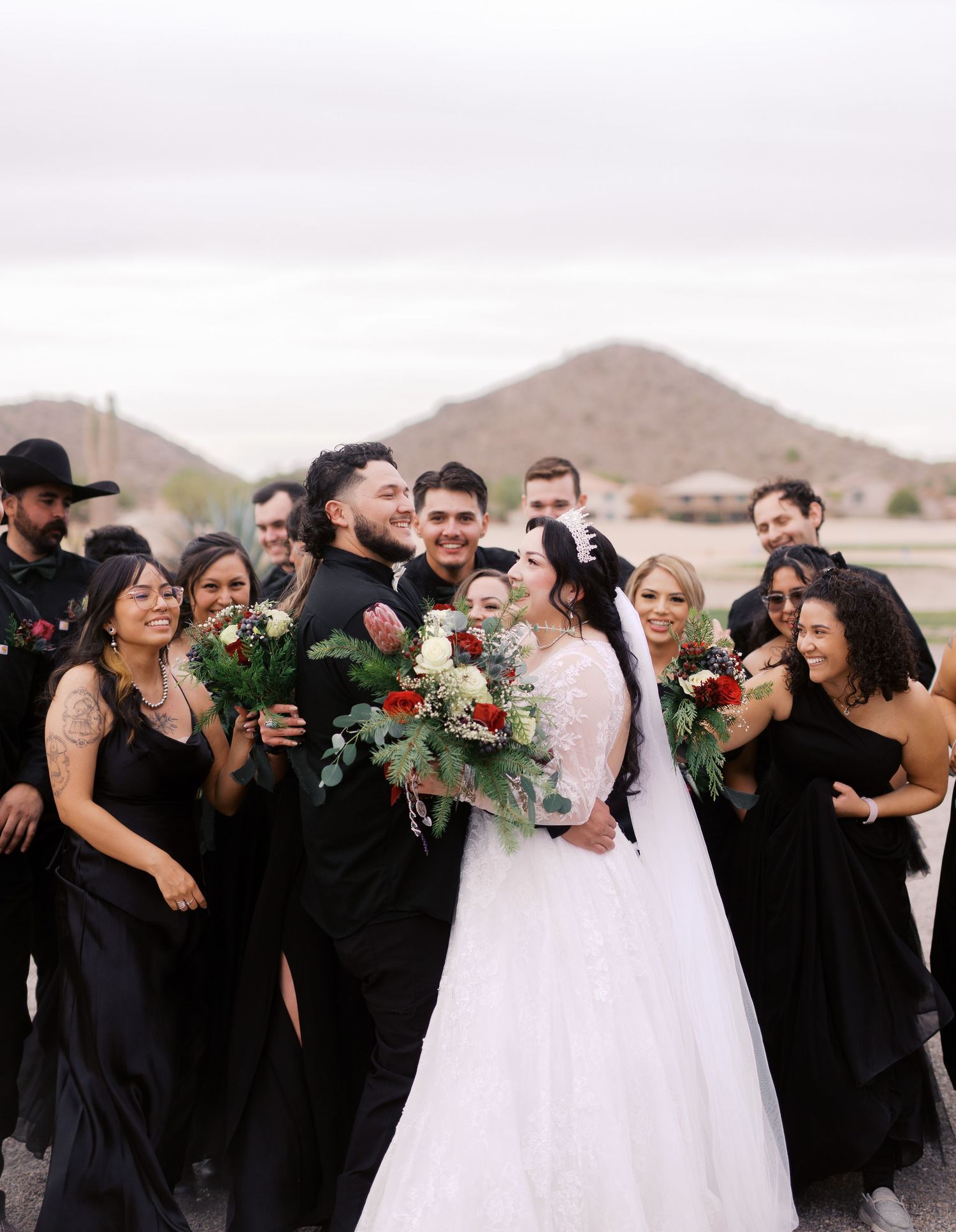 A wedding party laughs together outdoors in black attire with a desert mountain landscape in the background.