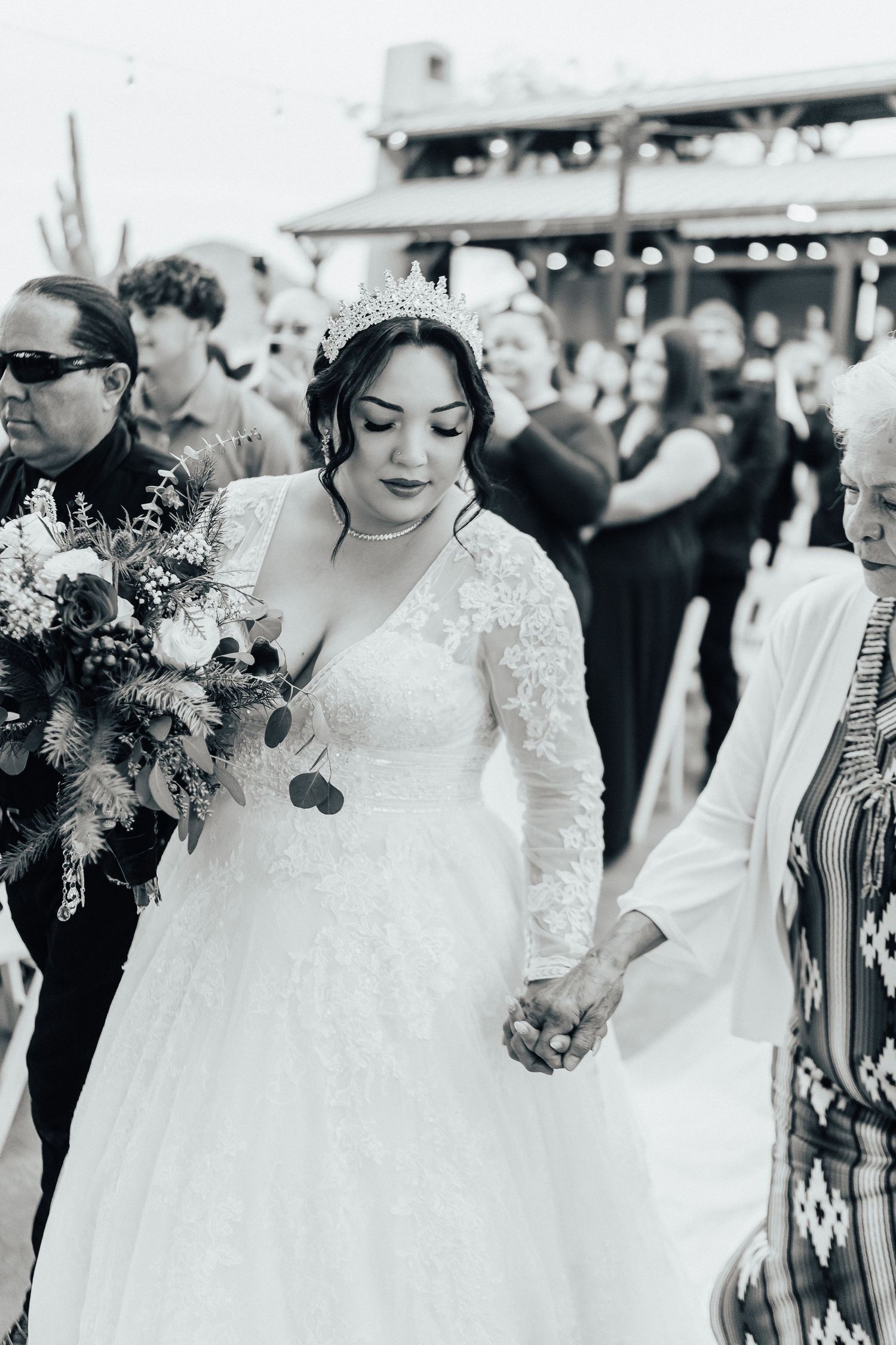 A bride in a flowing white lace gown holds a stunning wildflower bouquet at an outdoor wedding celebration.