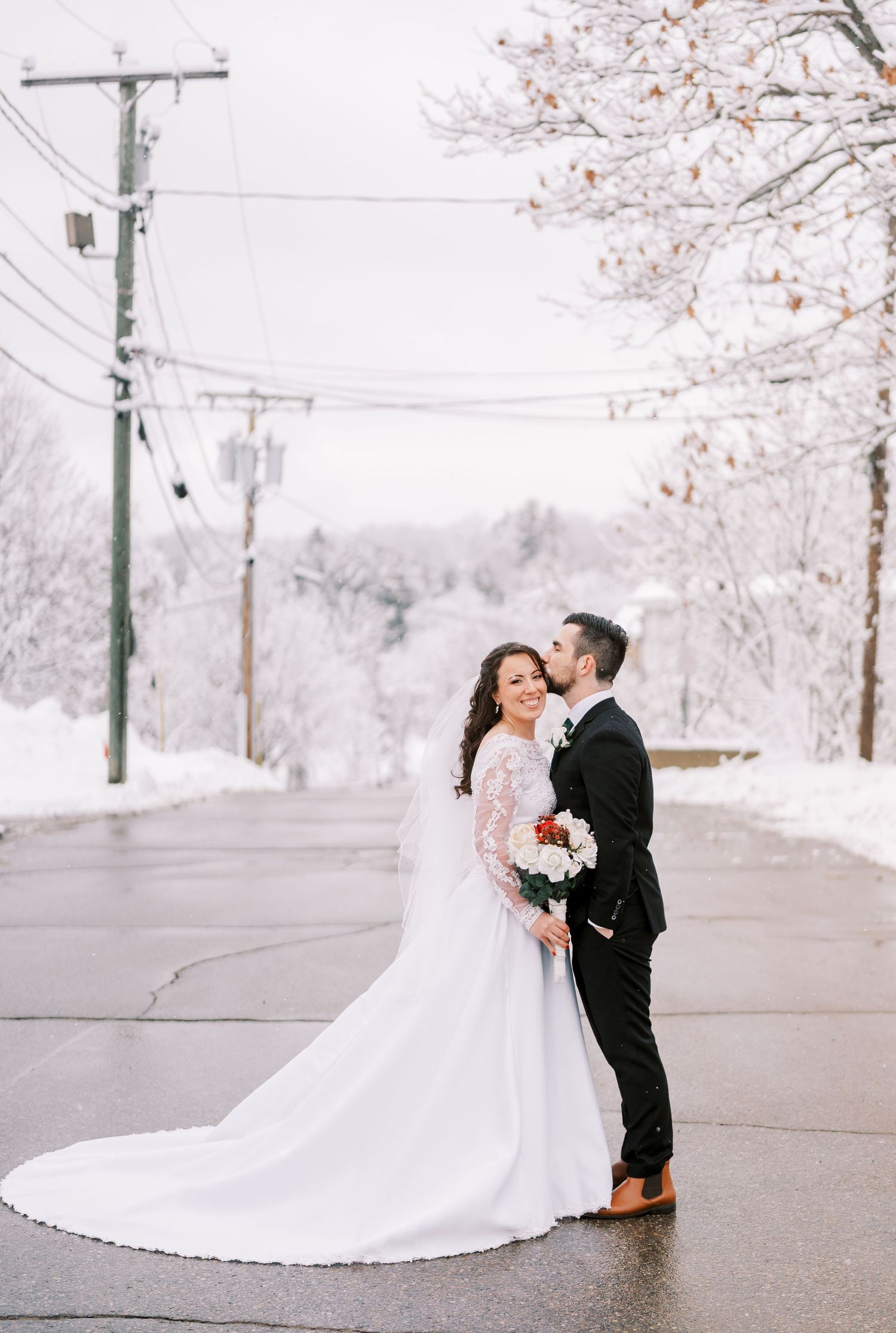 Newlyweds embrace on a snowy winter day with cherry blossom trees in the background.