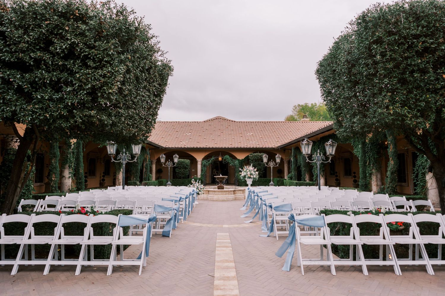 Outdoor wedding ceremony setup with white folding chairs and light blue cushions lined up beneath tall trees leading to wooden pavilion.