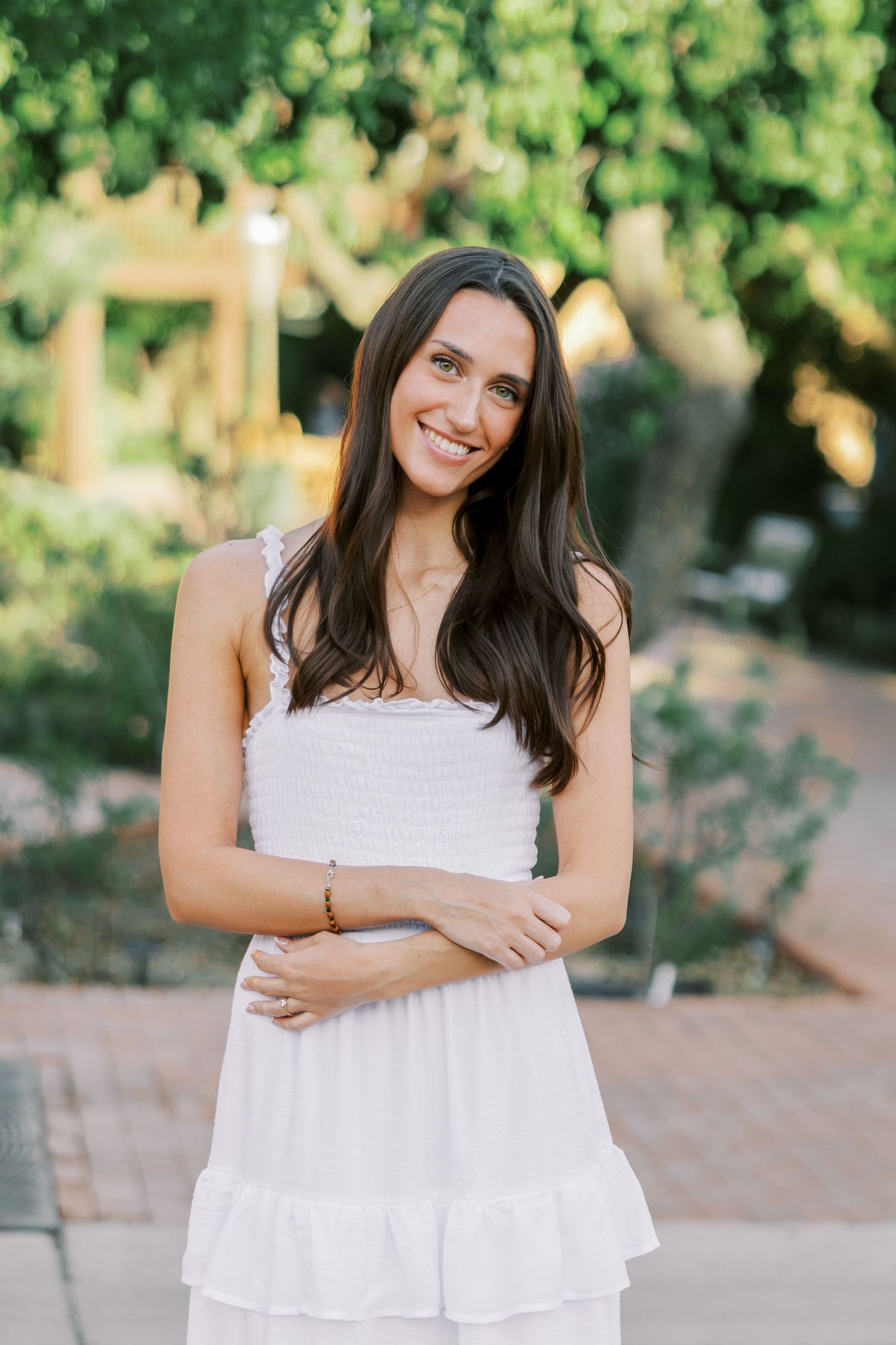 Woman in flowing white dress poses casually against lush green garden backdrop.