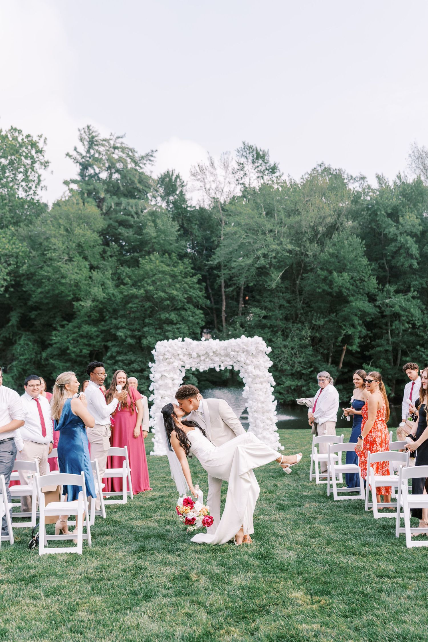 A romantic outdoor wedding ceremony with a white floral arch and guests seated on white chairs amid lush greenery.