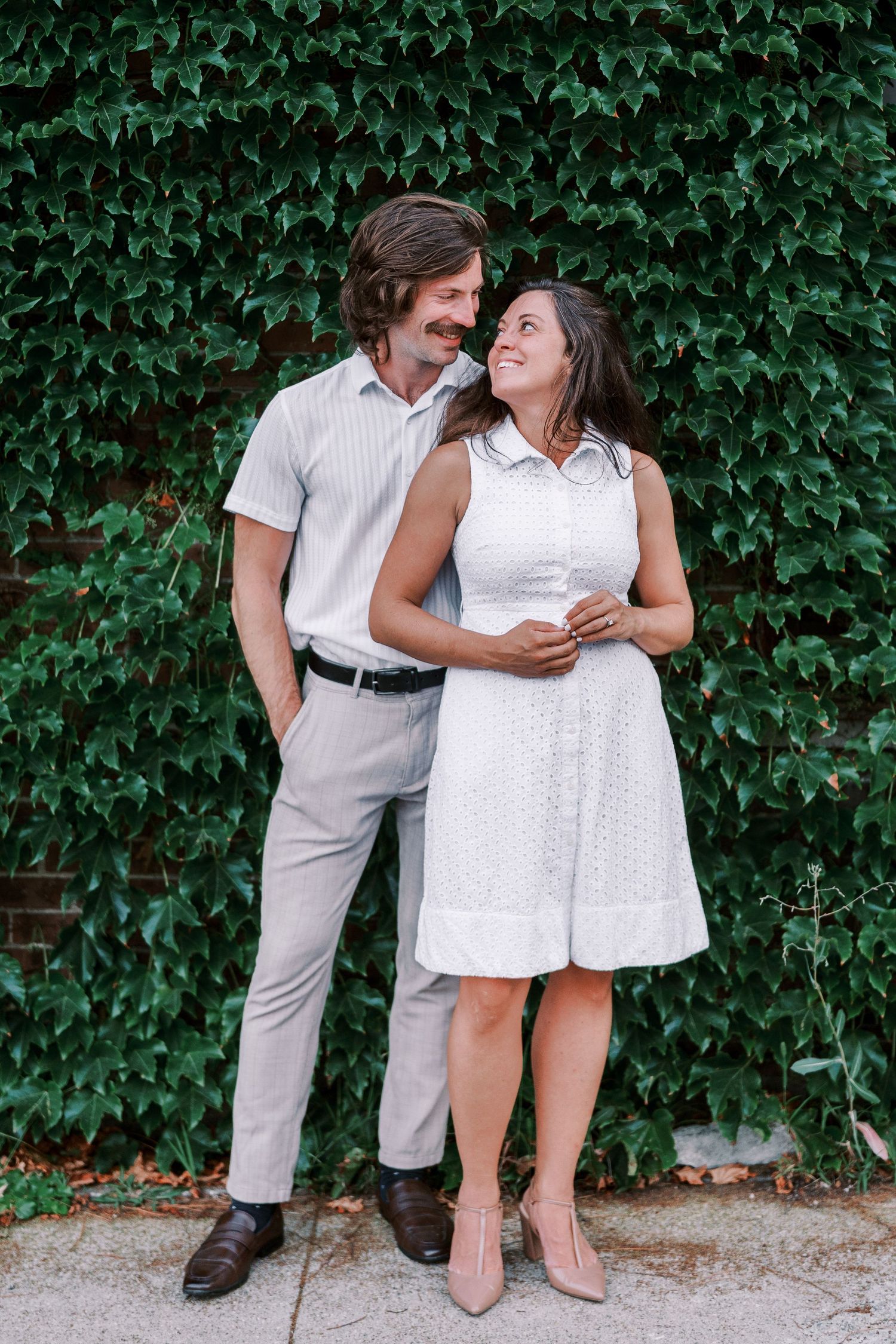 Dancing and laughing couple in white outfits pose against a lush green ivy wall.