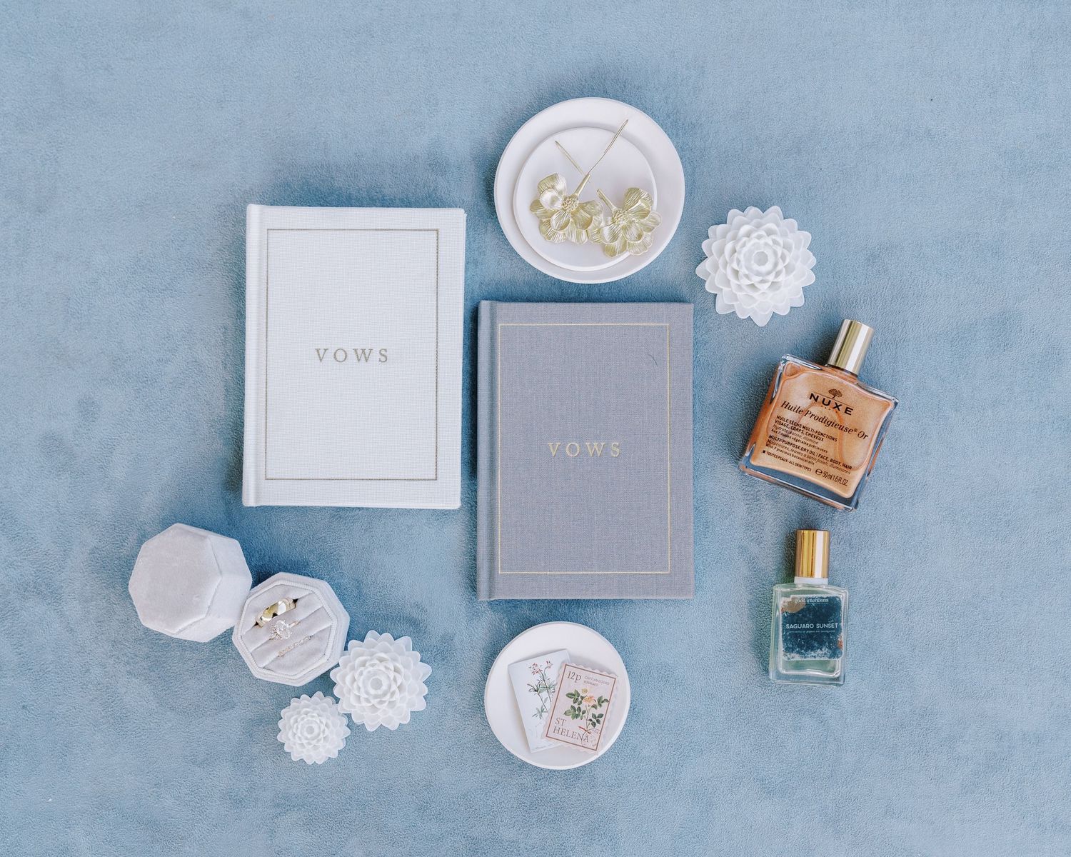 Flat lay of wedding accessories including perfume bottles and vow books on blue background.